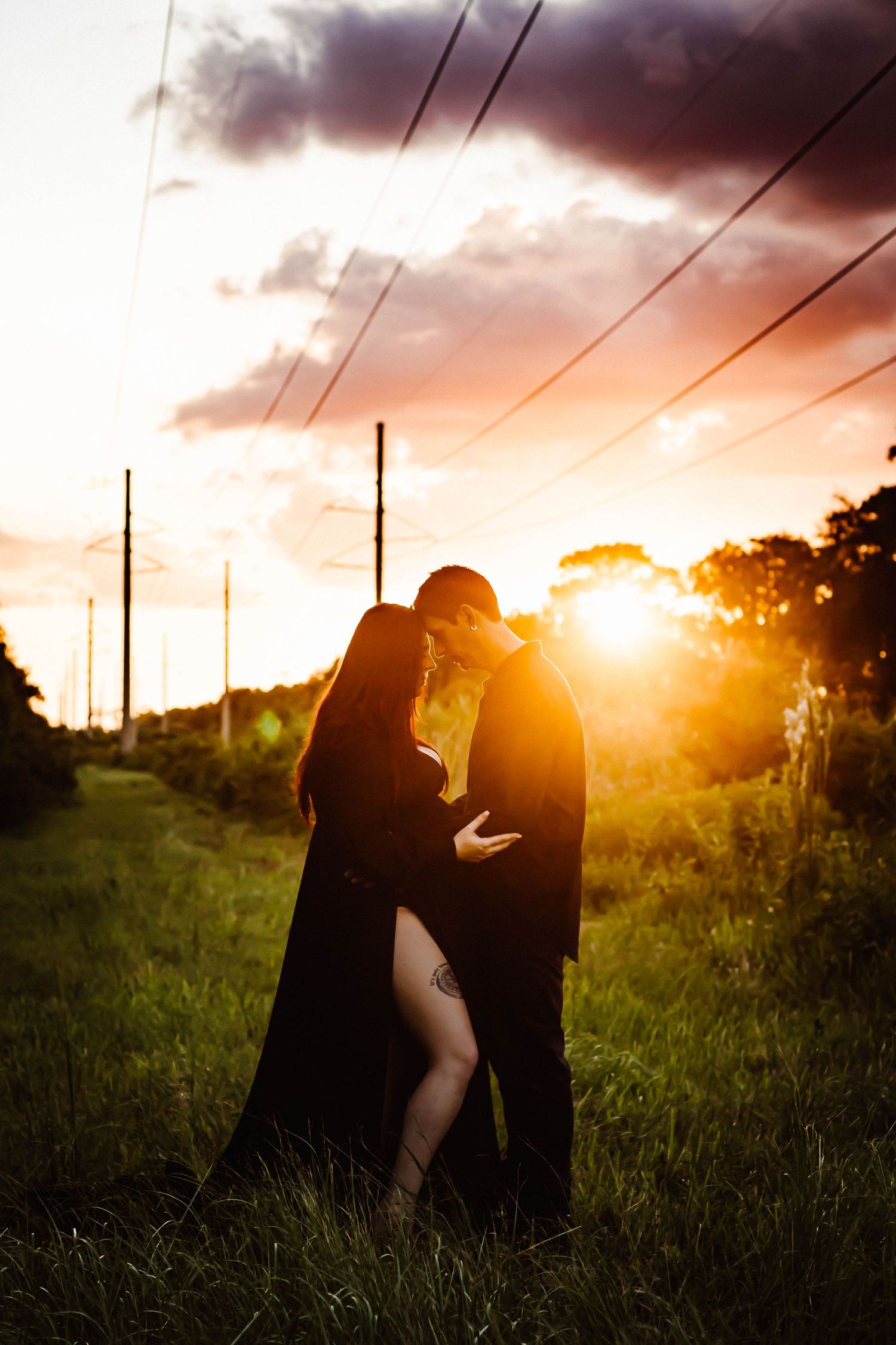 A romantic silhouette of a couple embracing in a field at sunset with dramatic clouds and power lines in the background.
