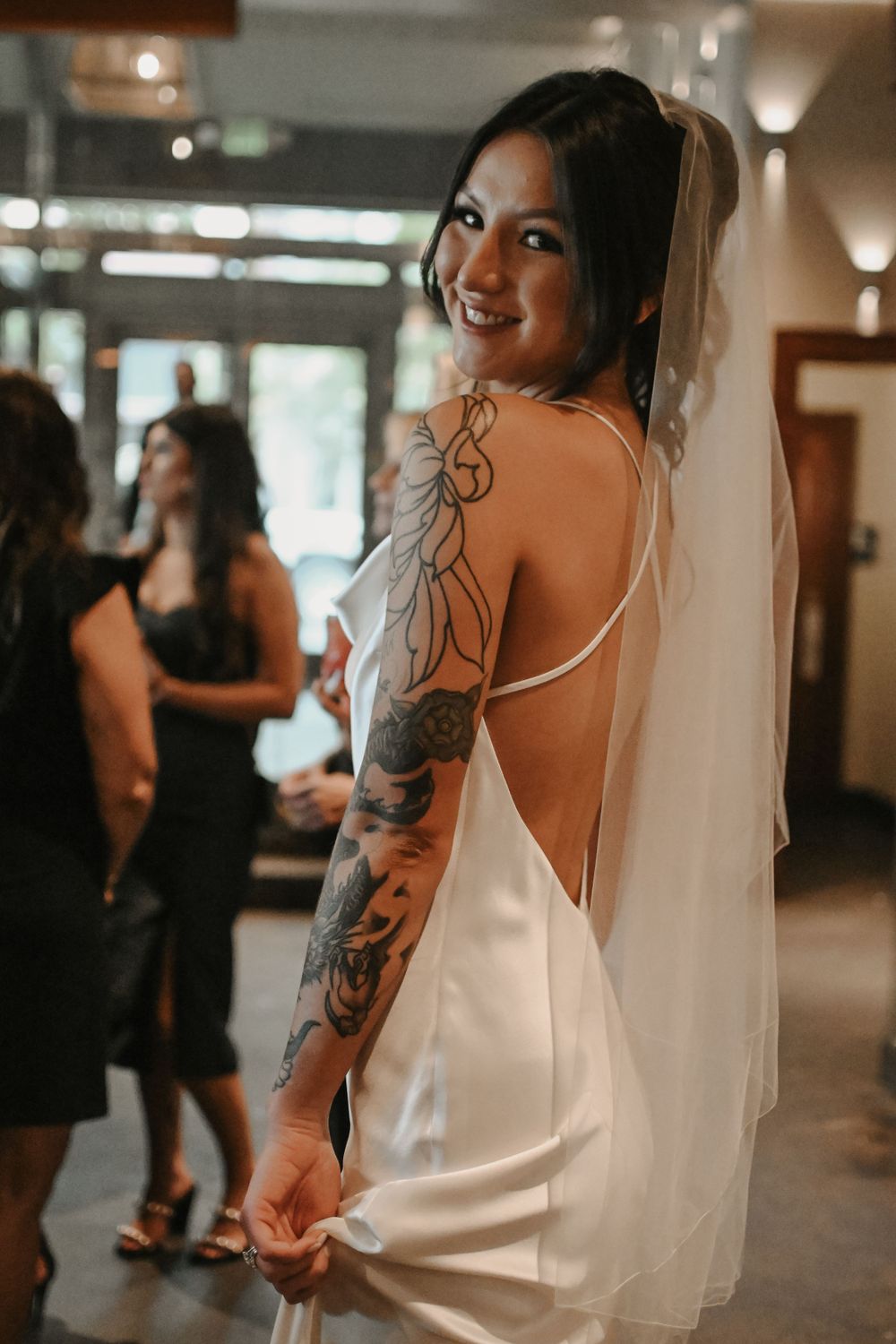 Bride in a white dress shows off artistic tattoo sleeves during wedding preparations.