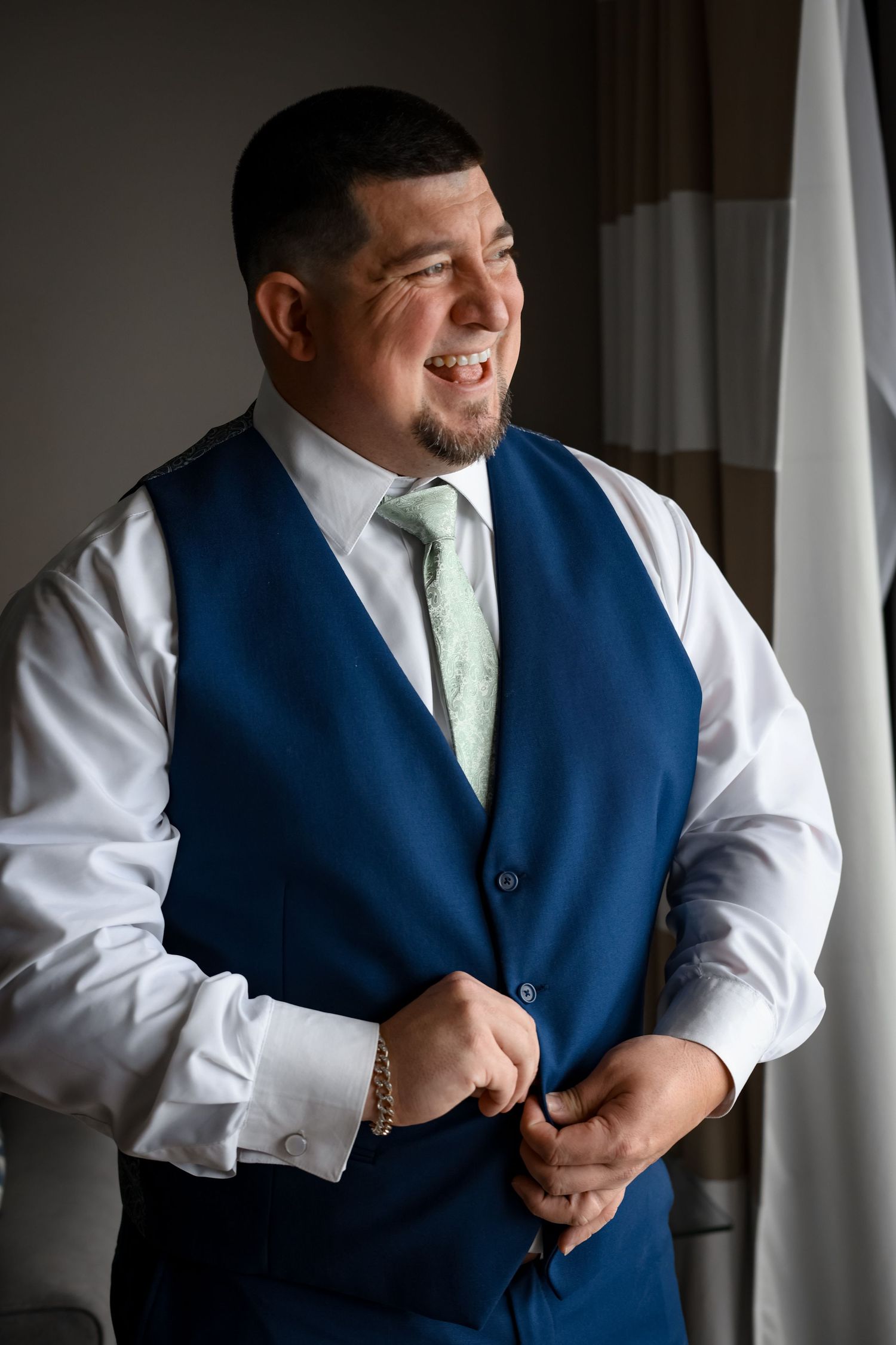 Groom in navy vest and mint tie smiles while getting ready.