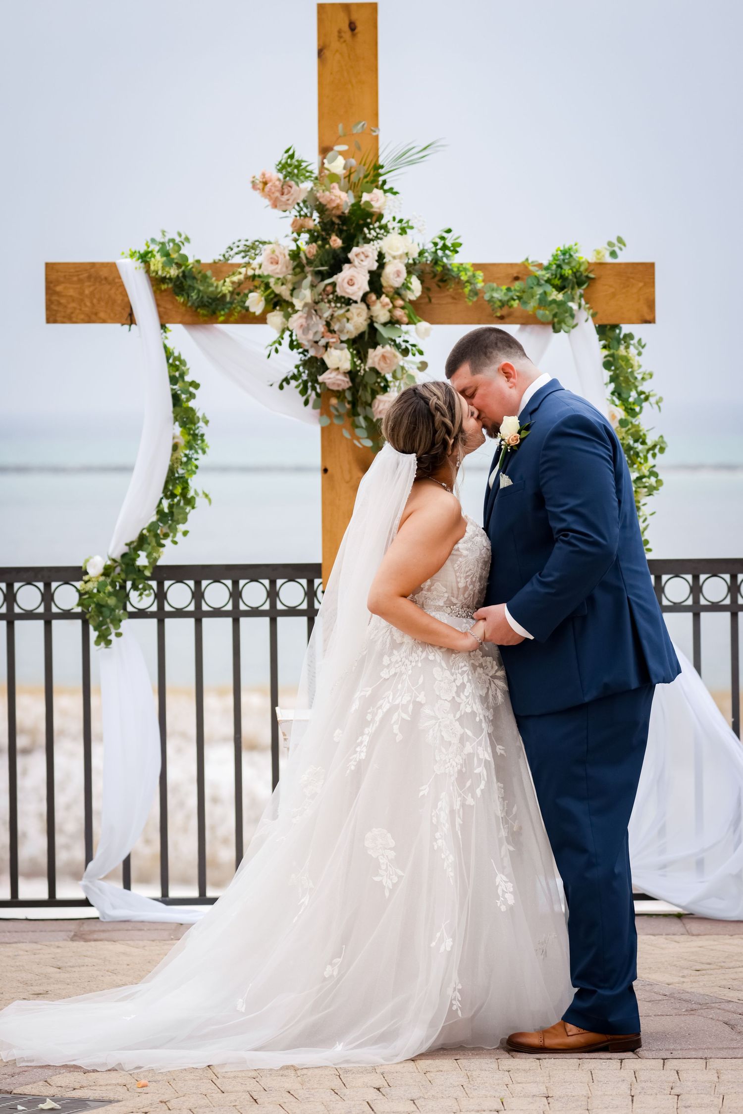 Wedding couple shares intimate moment under floral cross arch by the ocean.