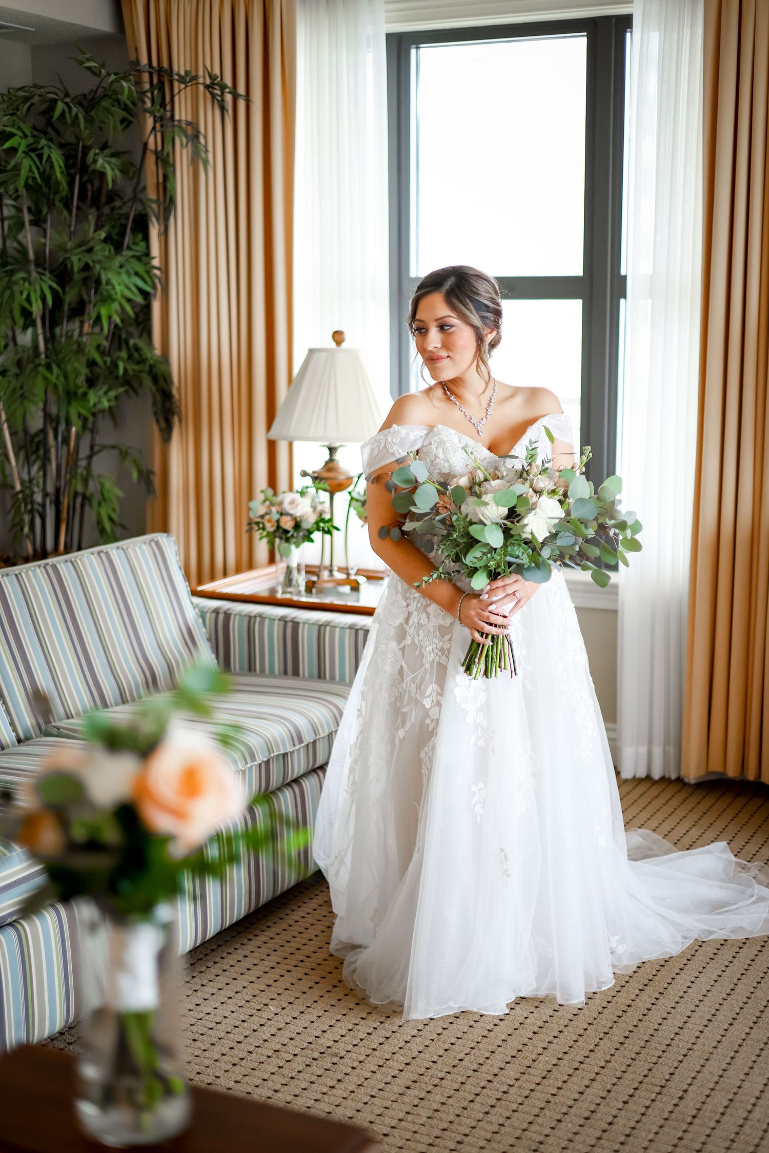 Bride in white lace gown holds elegant white and green bouquet near window.