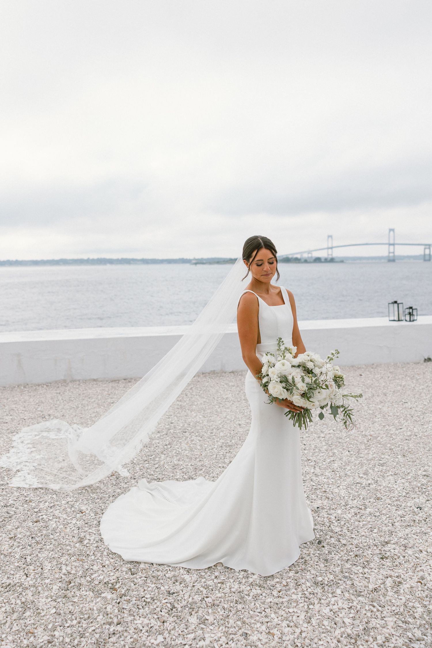 A flowing white wedding dress trails elegantly across a pebbly beach with water views.