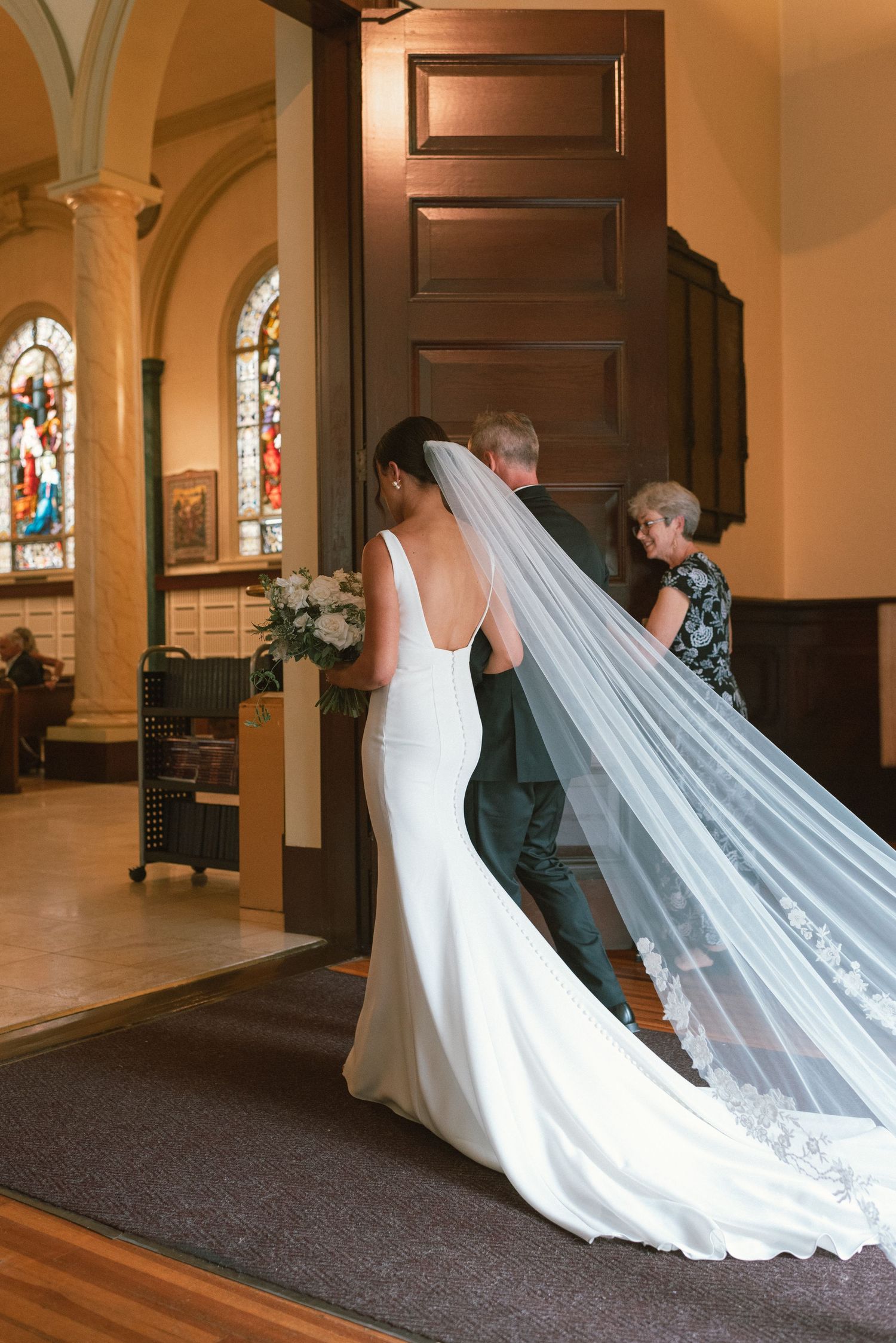 Long flowing wedding veil trails behind bride as she walks through church interior with stained glass windows.