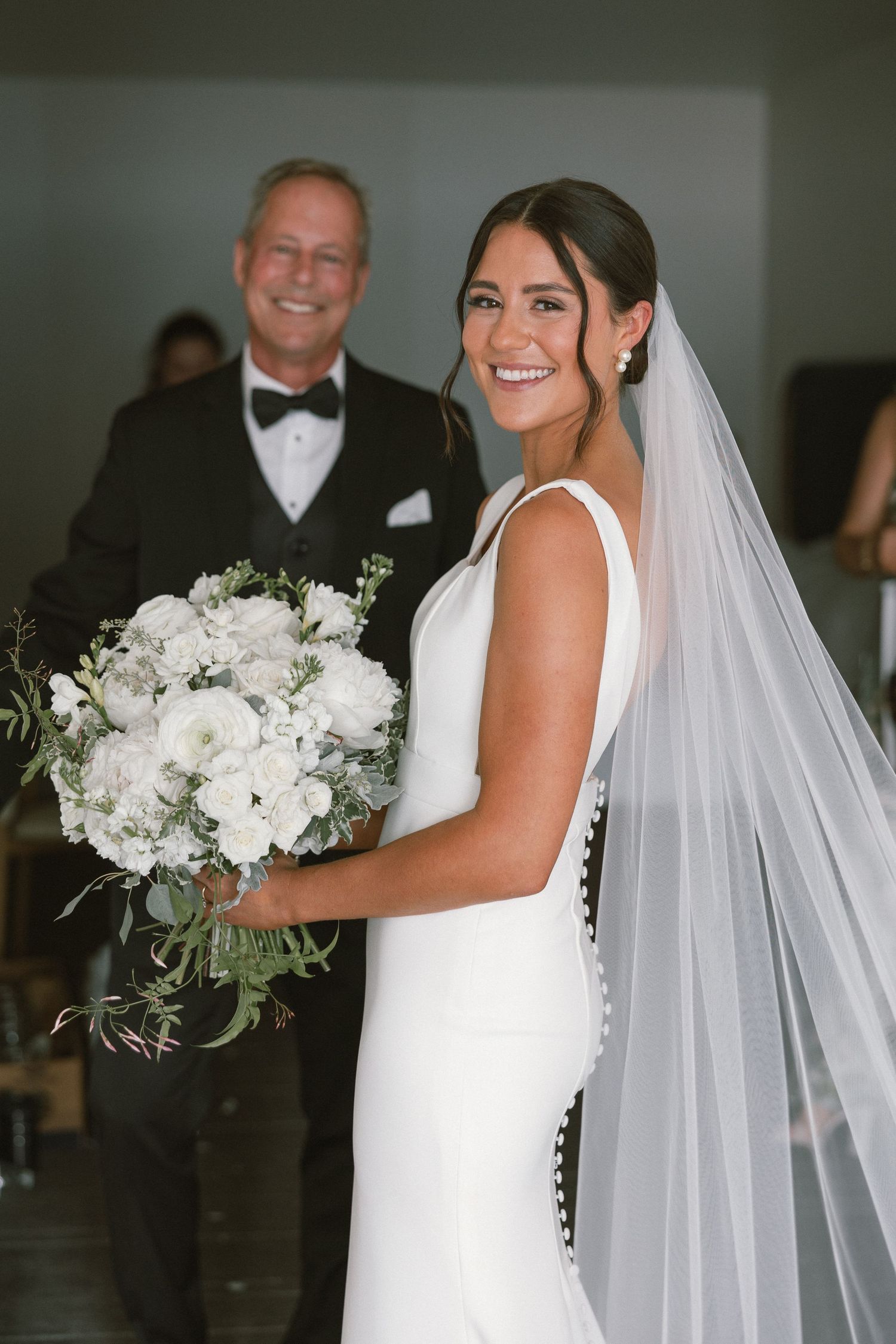 Bride in a sleeveless white wedding dress holds a lush white bouquet while wearing a flowing cathedral veil.