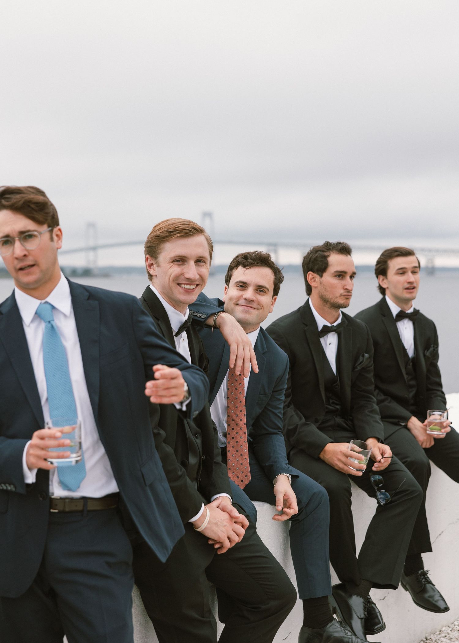 Groomsmen in dark suits sit casually on beach wall during wedding photoshoot.