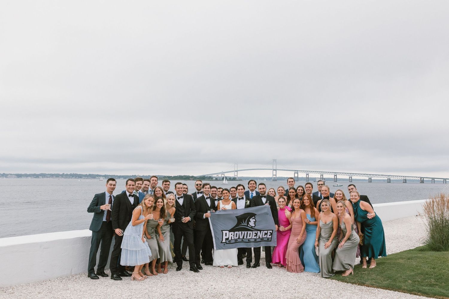 Large wedding party poses together on beach with bridge visible in background.
