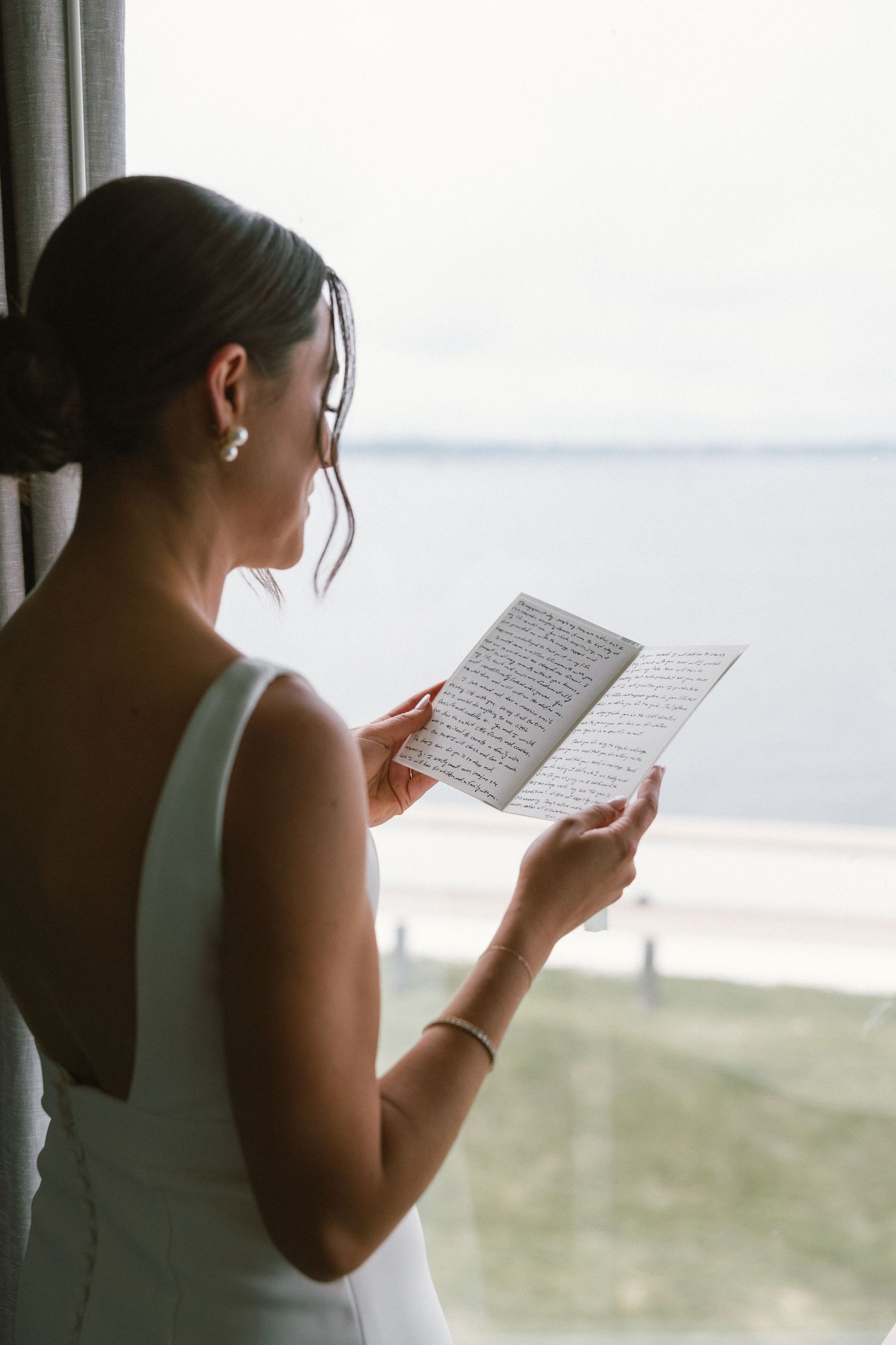 Person in white dress reading handwritten notes by a window overlooking the ocean.