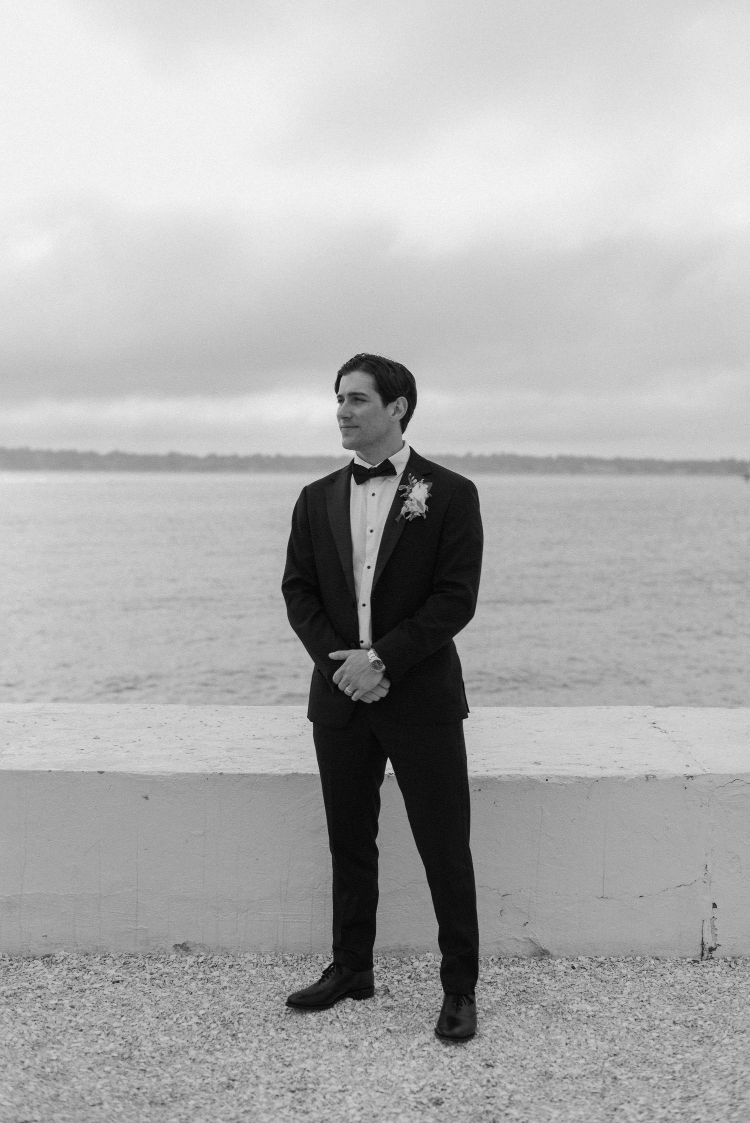 Groomsmen in classic black tuxedos pose casually on a rocky beach.