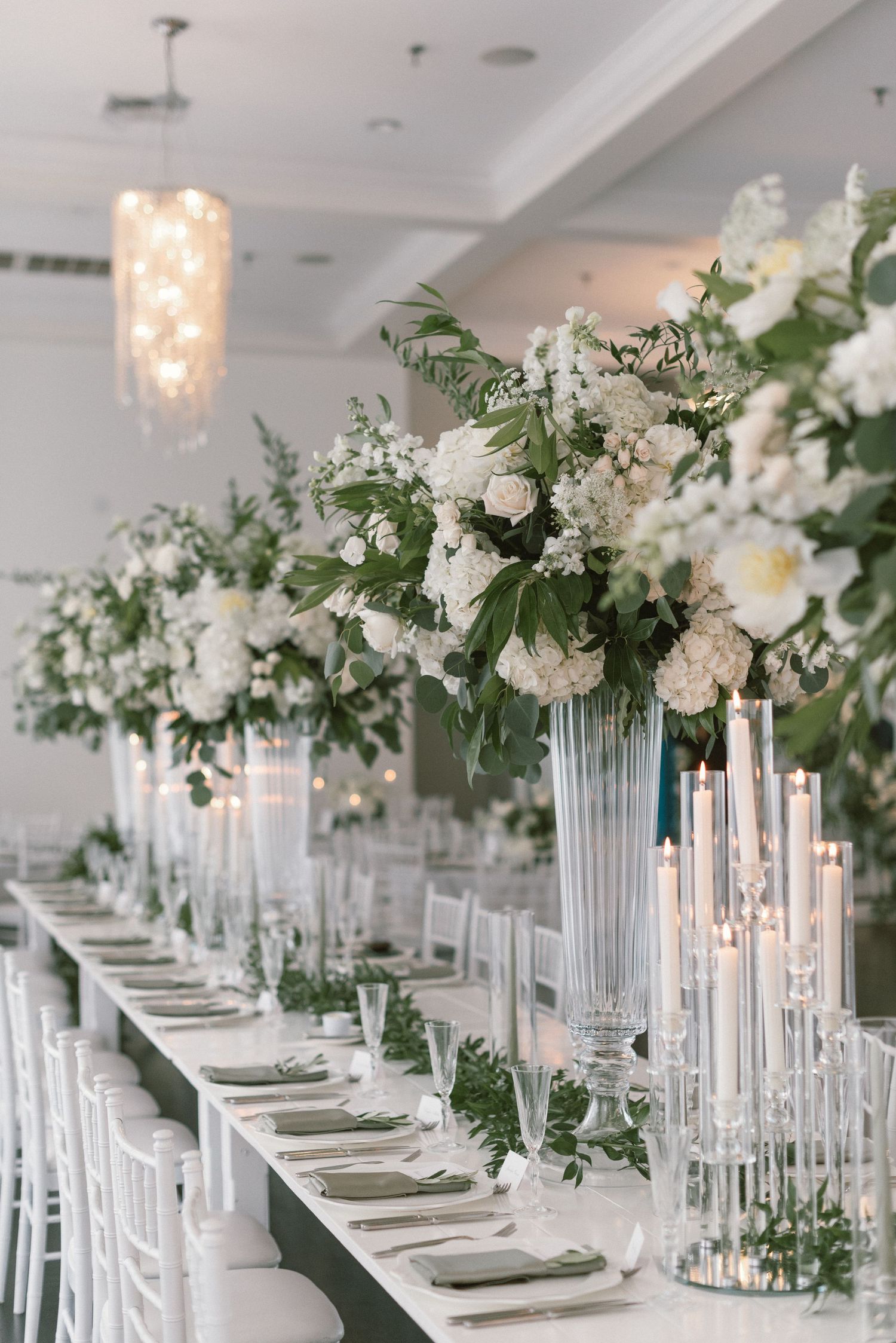 Elegant wedding reception table with tall glass vases, white flowers and crystal chandeliers.