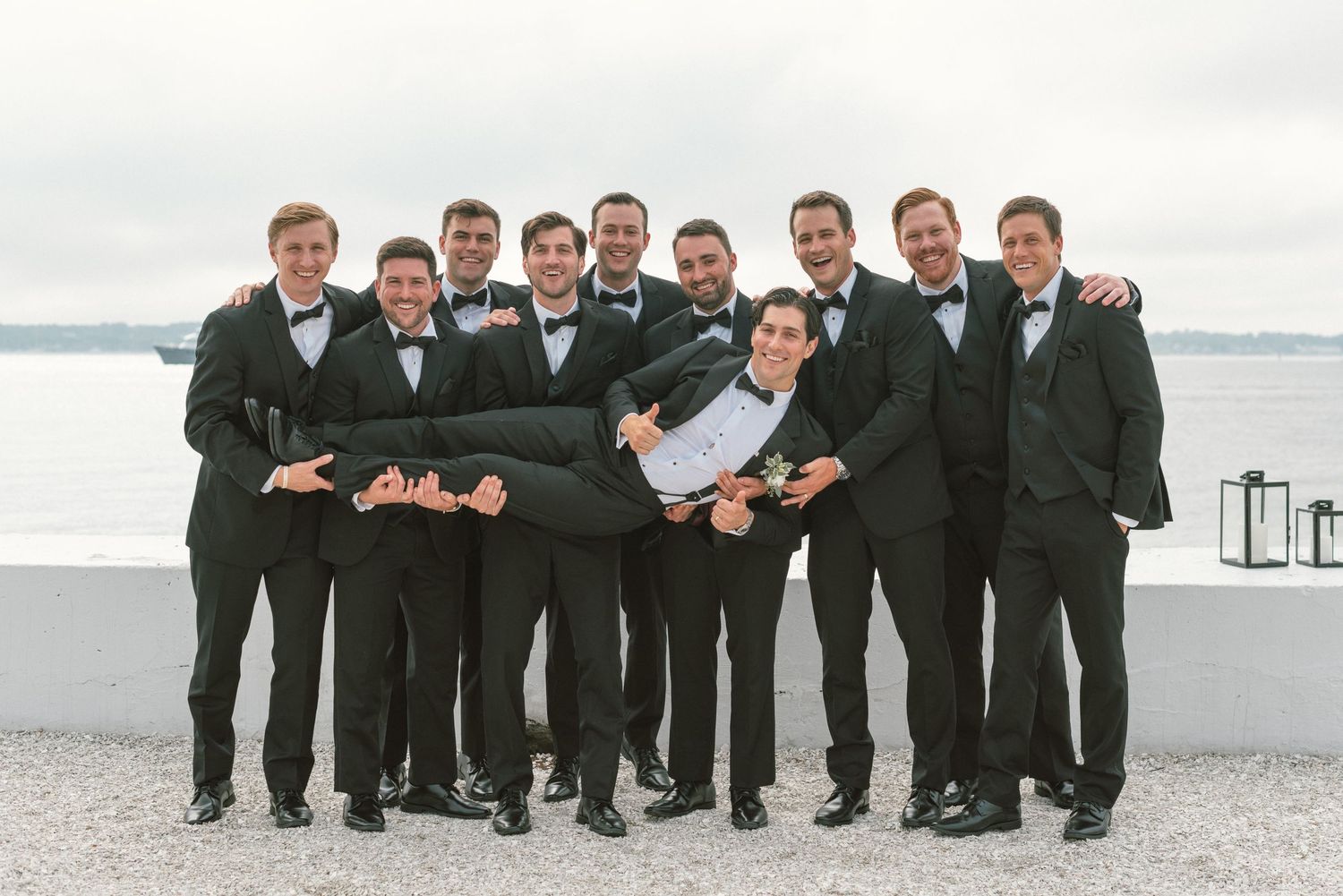 Groomsmen in black tuxedos pose playfully on a beach with water and bridge in background.