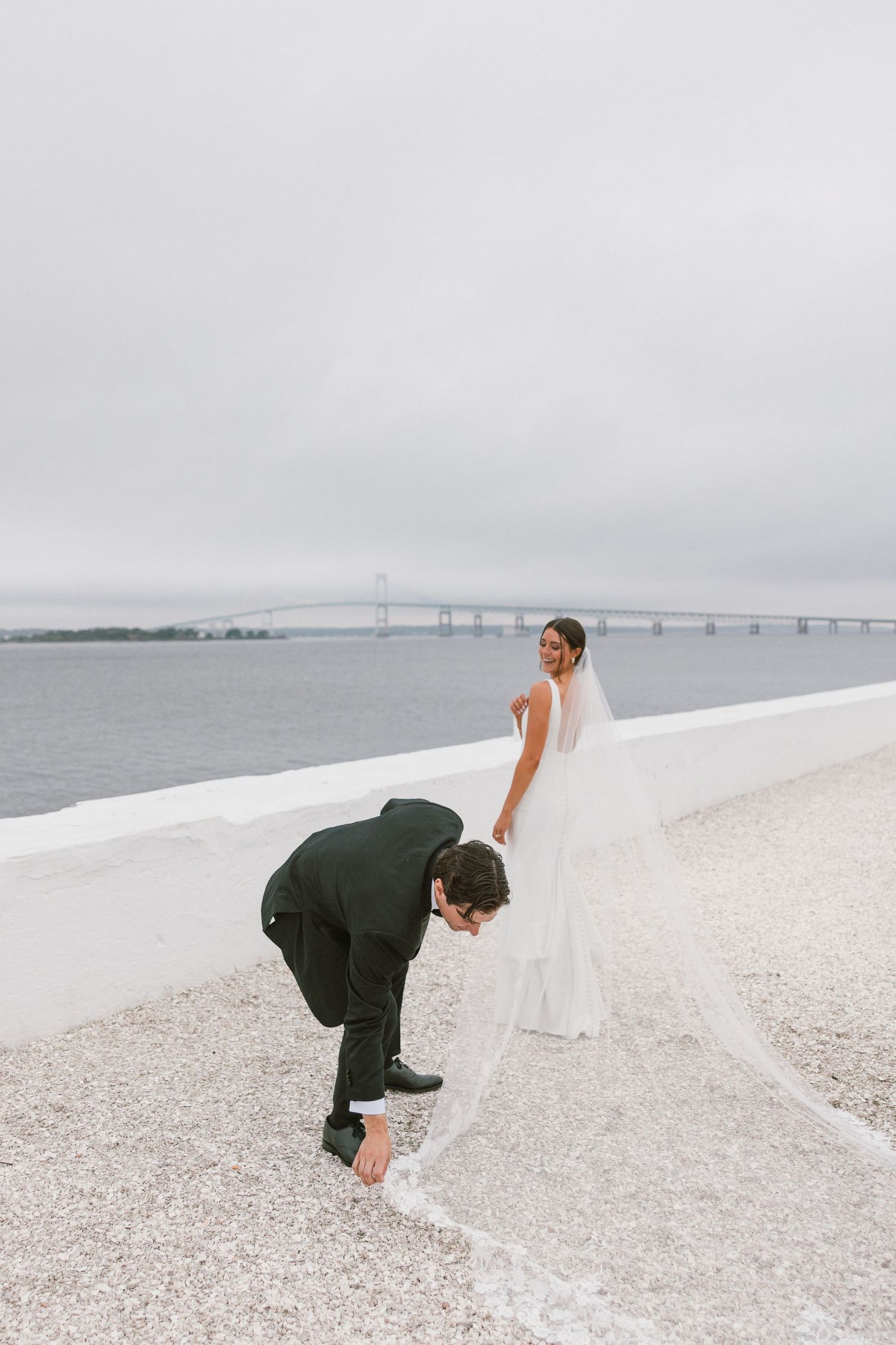 Wedding couple playing on a white sandy beach with a bridge visible in the background.