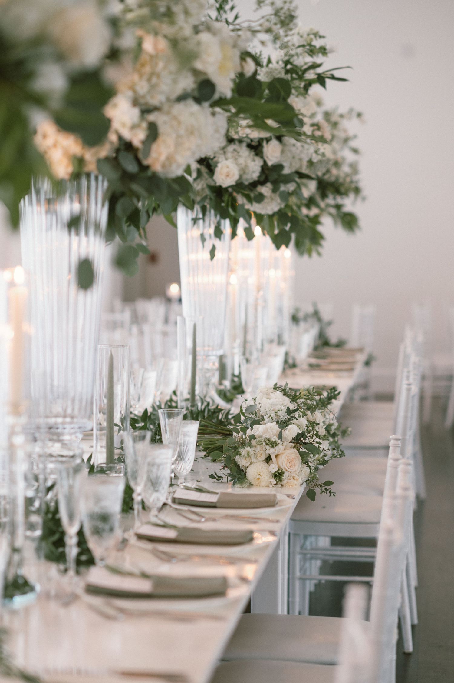 Elegant wedding reception table settings with white floral centerpieces and crystal candle holders in a light-filled venue.