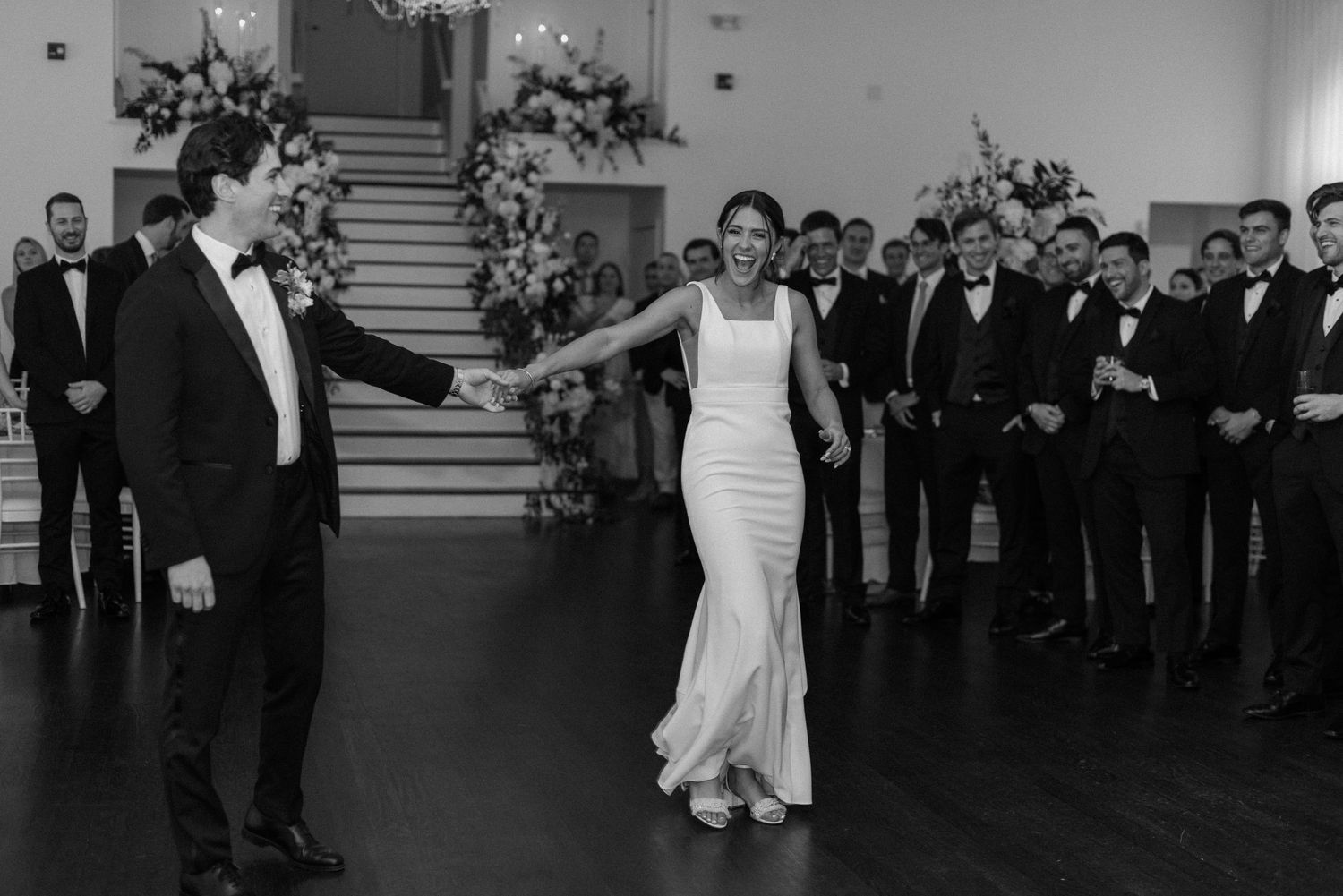 Wedding party dancing and celebrating in a spacious ballroom with elegant decor.