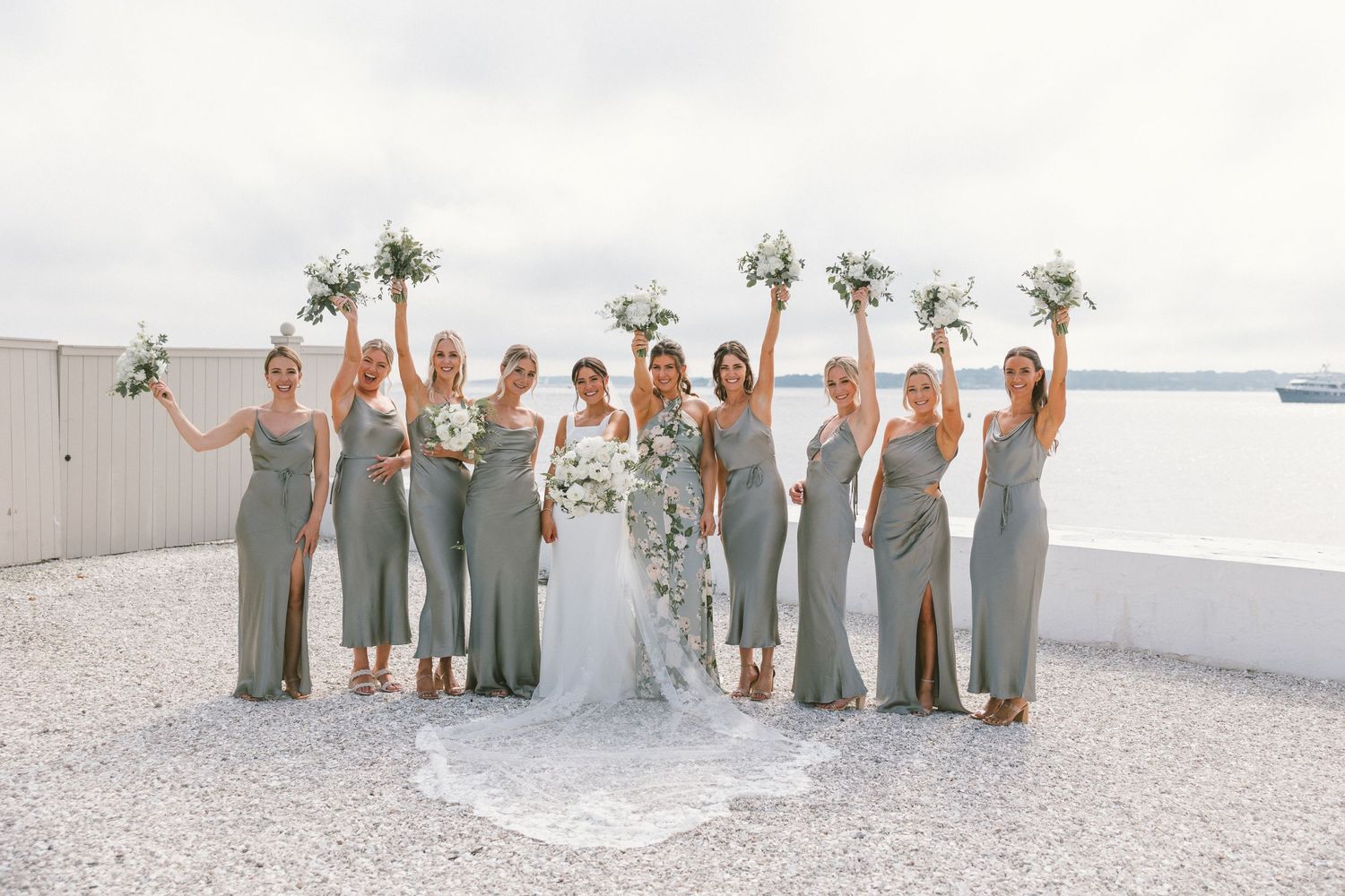 Bridesmaids in sage green dresses pose with the bride on a sunny beach.