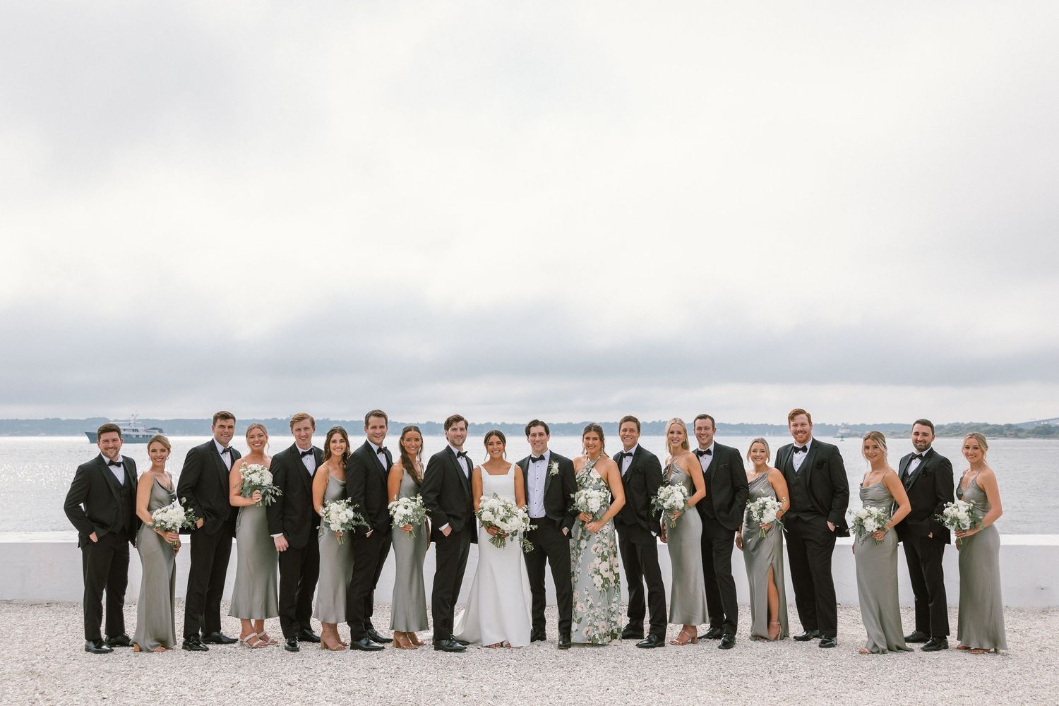 Wedding party lines up for formal portrait on beach with bridesmaids in sage and groomsmen in black.