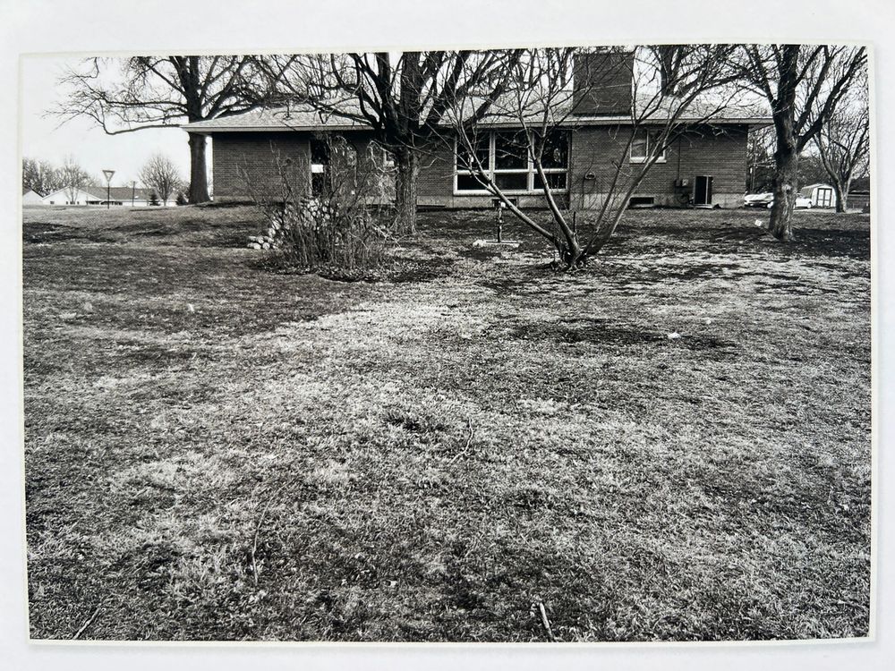 Black and white landscape view of an empty field with trees and buildings in background.