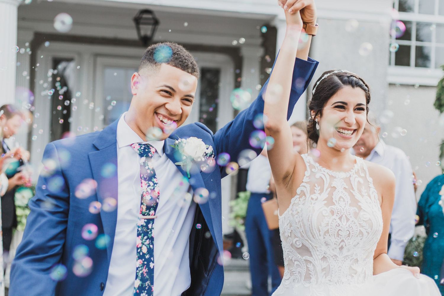Joyful newlyweds celebrate with raised arms as bubbles float around them outside a building.