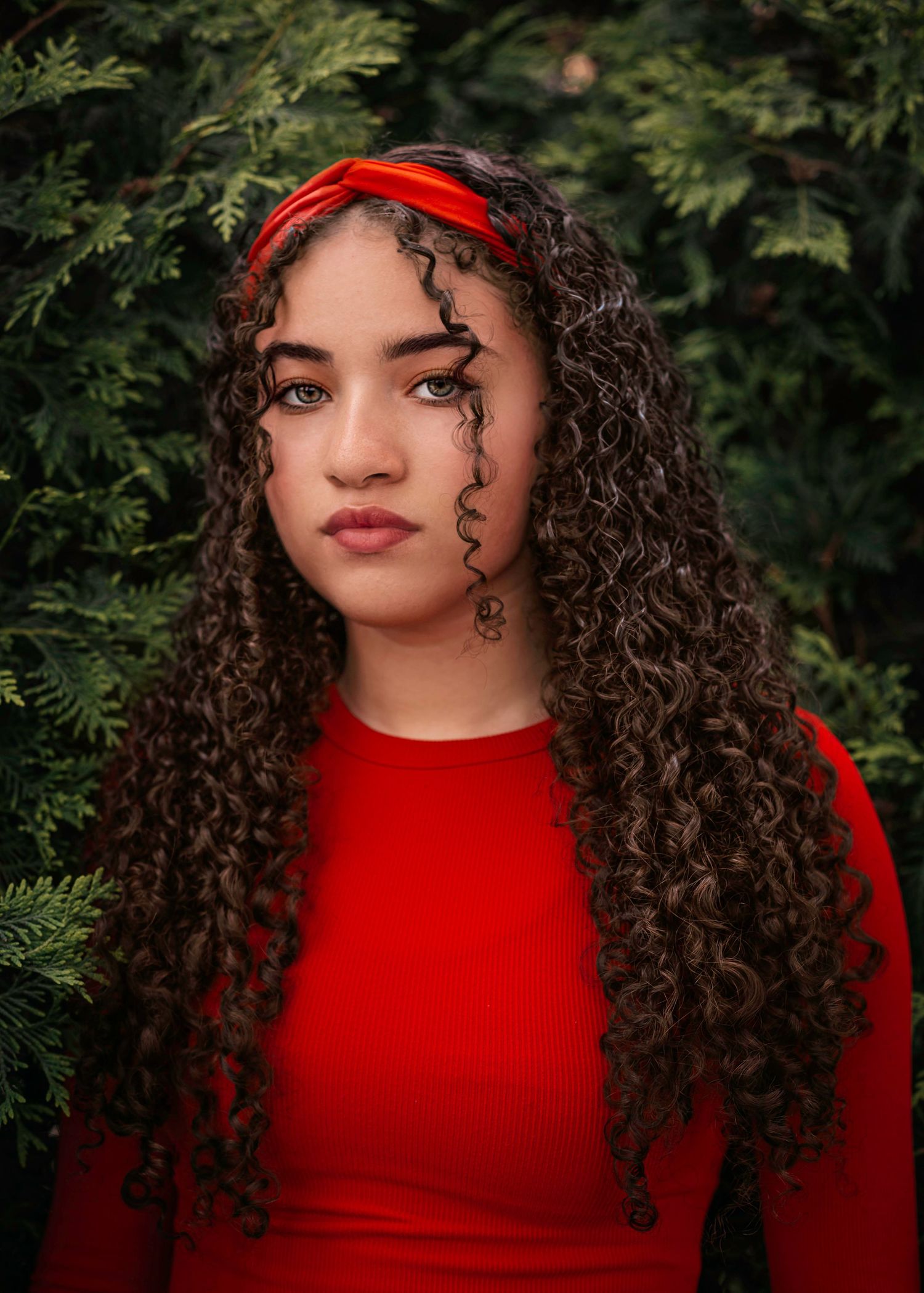 Close-up portrait with red headband and matching top against lush green foliage.