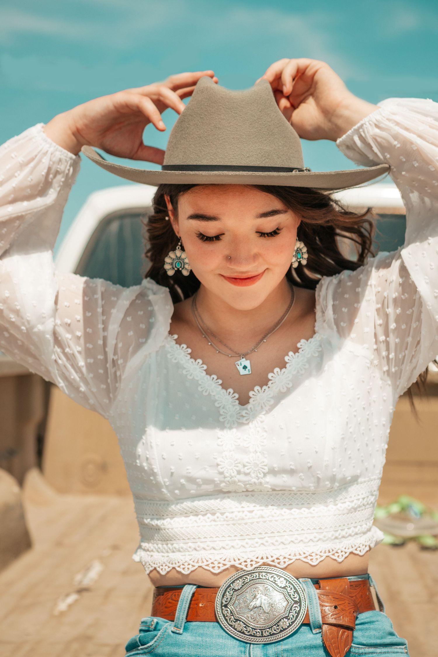 Western style portrait in white lace top and gray cowboy hat with ornate belt buckle.