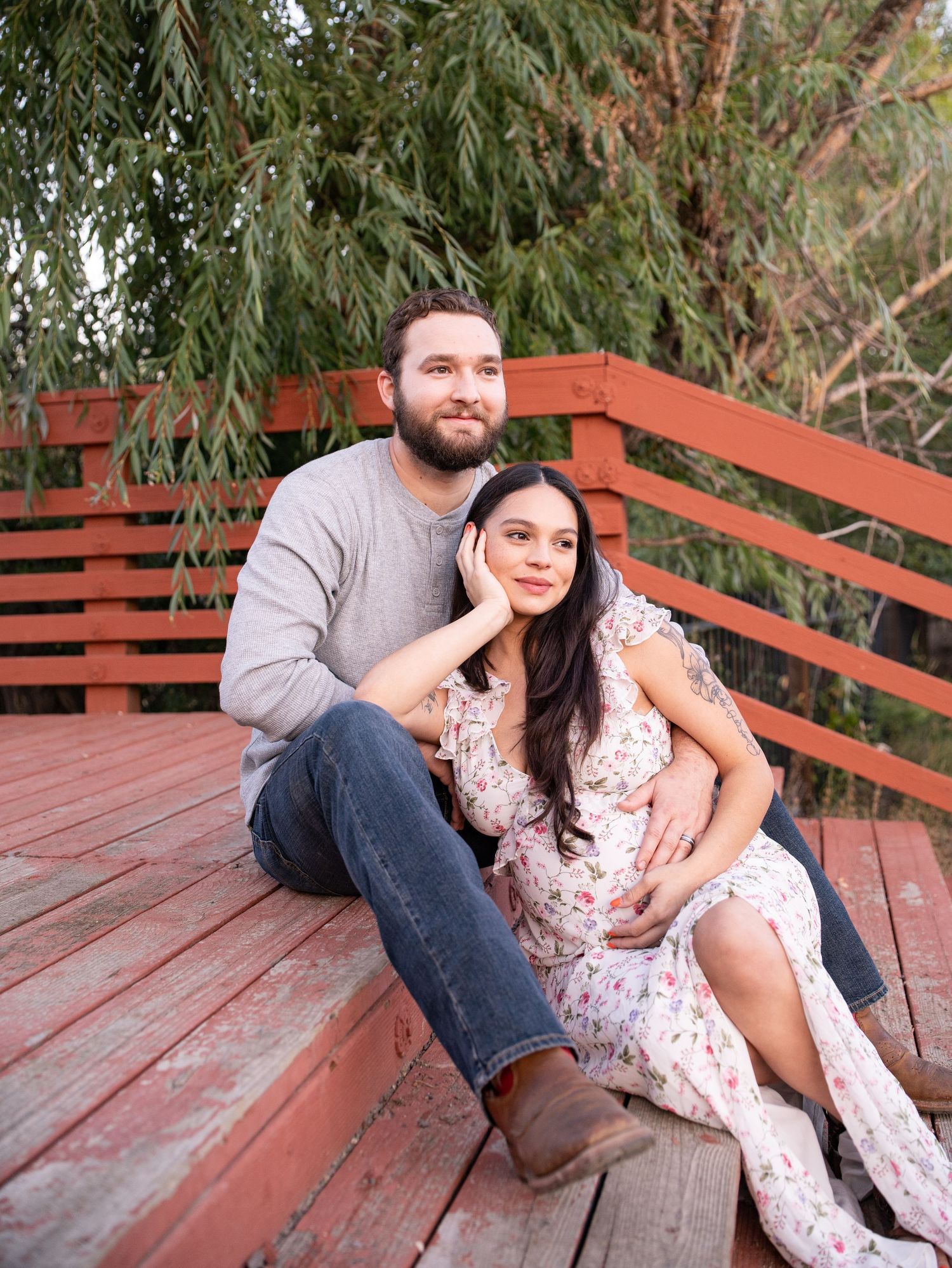 Couple sitting together on wooden deck stairs with trees in background during outdoor maternity photo session.