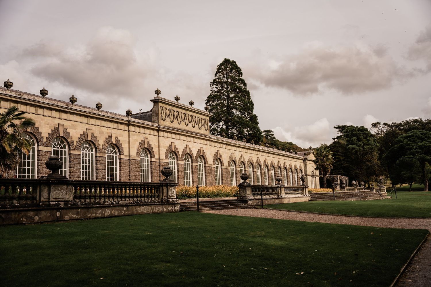 Grand classical orangery building with ornate architecture and landscaped grounds under moody skies.