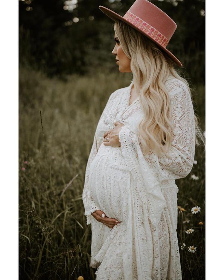 Bohemian maternity portrait of woman in white lace dress and pink hat in meadow.