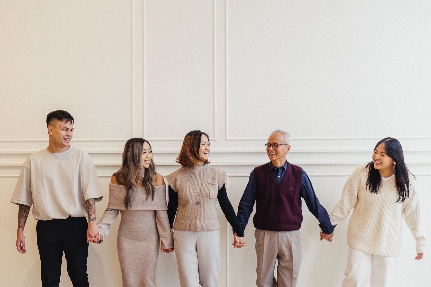 People holding hands in a line against a white wall wearing coordinated neutral tones.