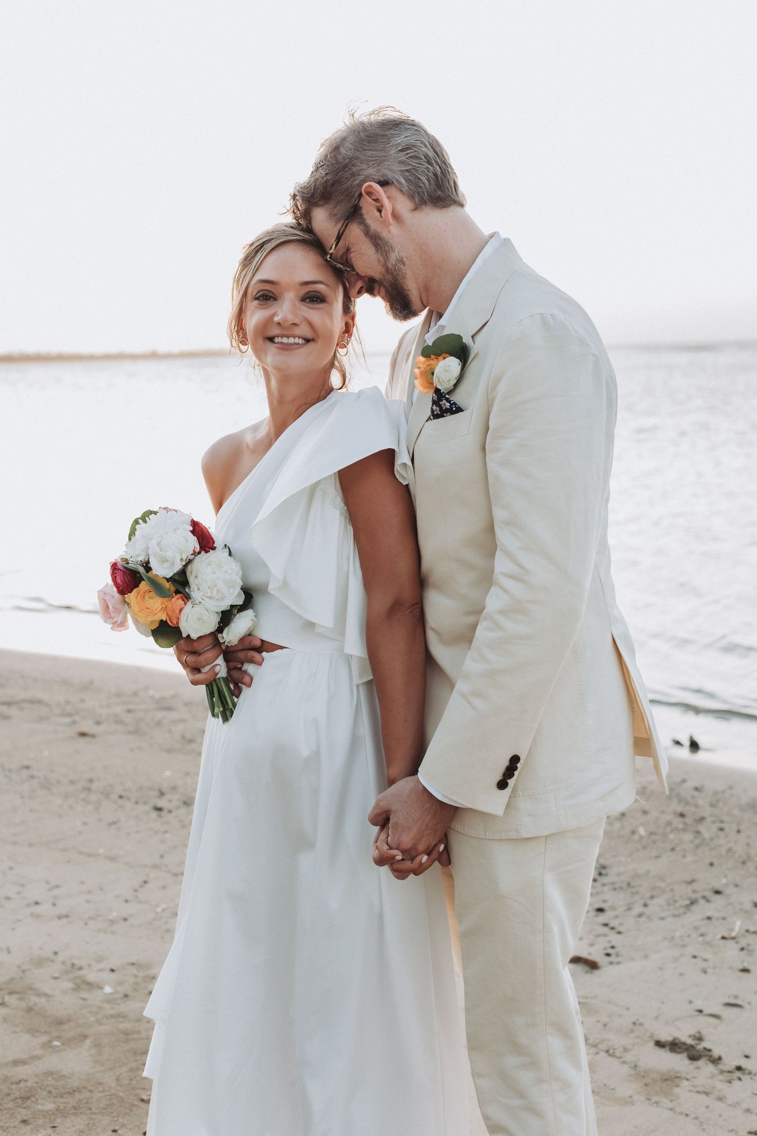 Couple in white wedding attire embracing on beach at sunset.