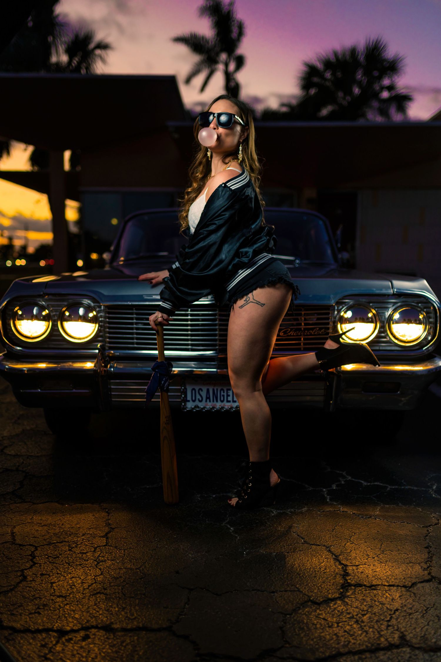 Dramatic nighttime photo with vintage car headlights glowing against a purple sunset sky and palm tree silhouettes.