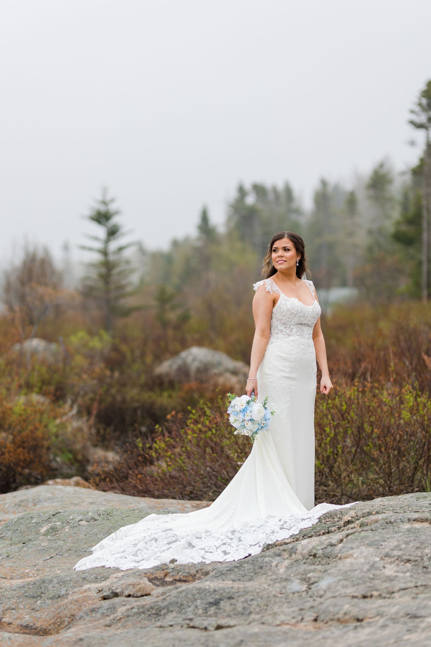 Elegant bride in lace gown holding blue bouquet poses on rocky terrain with forest backdrop.