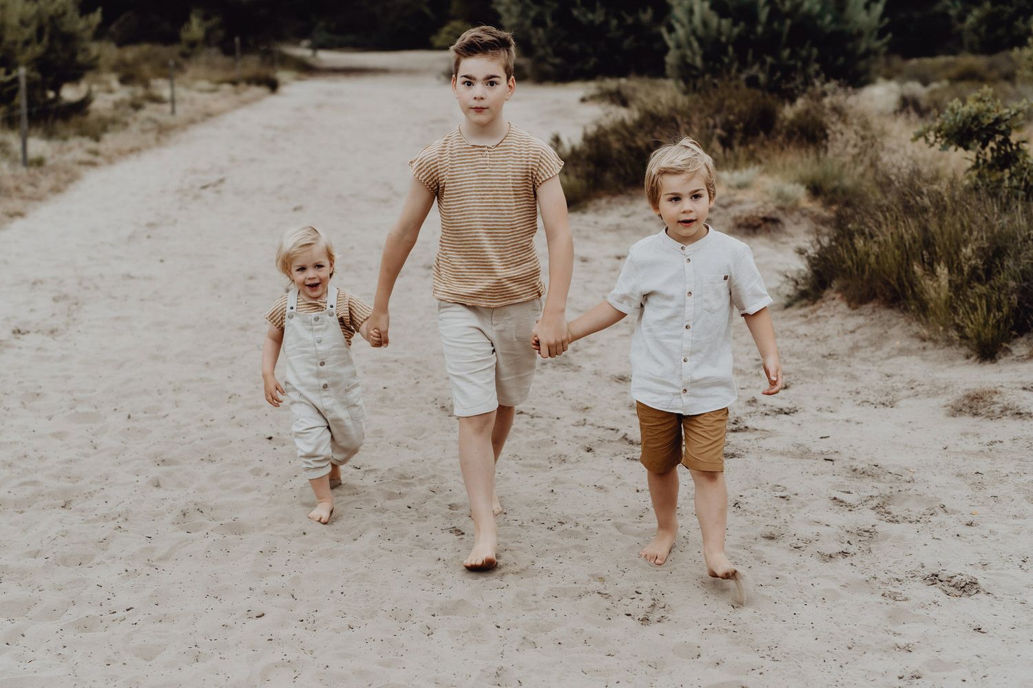 Three children walking hand in hand on sandy path in muted earth tones.
