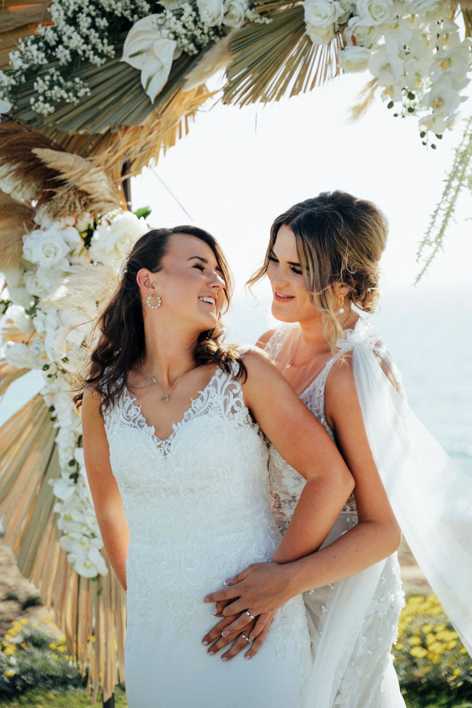 Two happy people in white wedding dresses share an intimate moment under a floral arch with palm fronds.