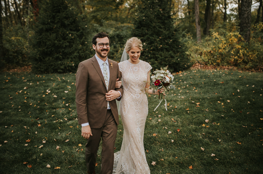A couple standing together on grass with fall foliage holding a wedding bouquet.