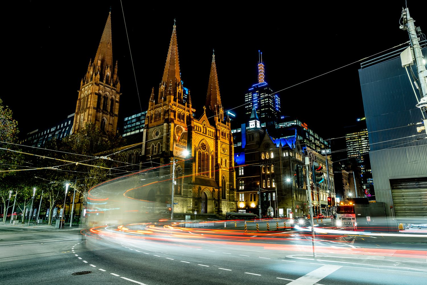 St Pauls Cathedral Melbourne illuminated at night with light trails from passing trams on busy intersection.