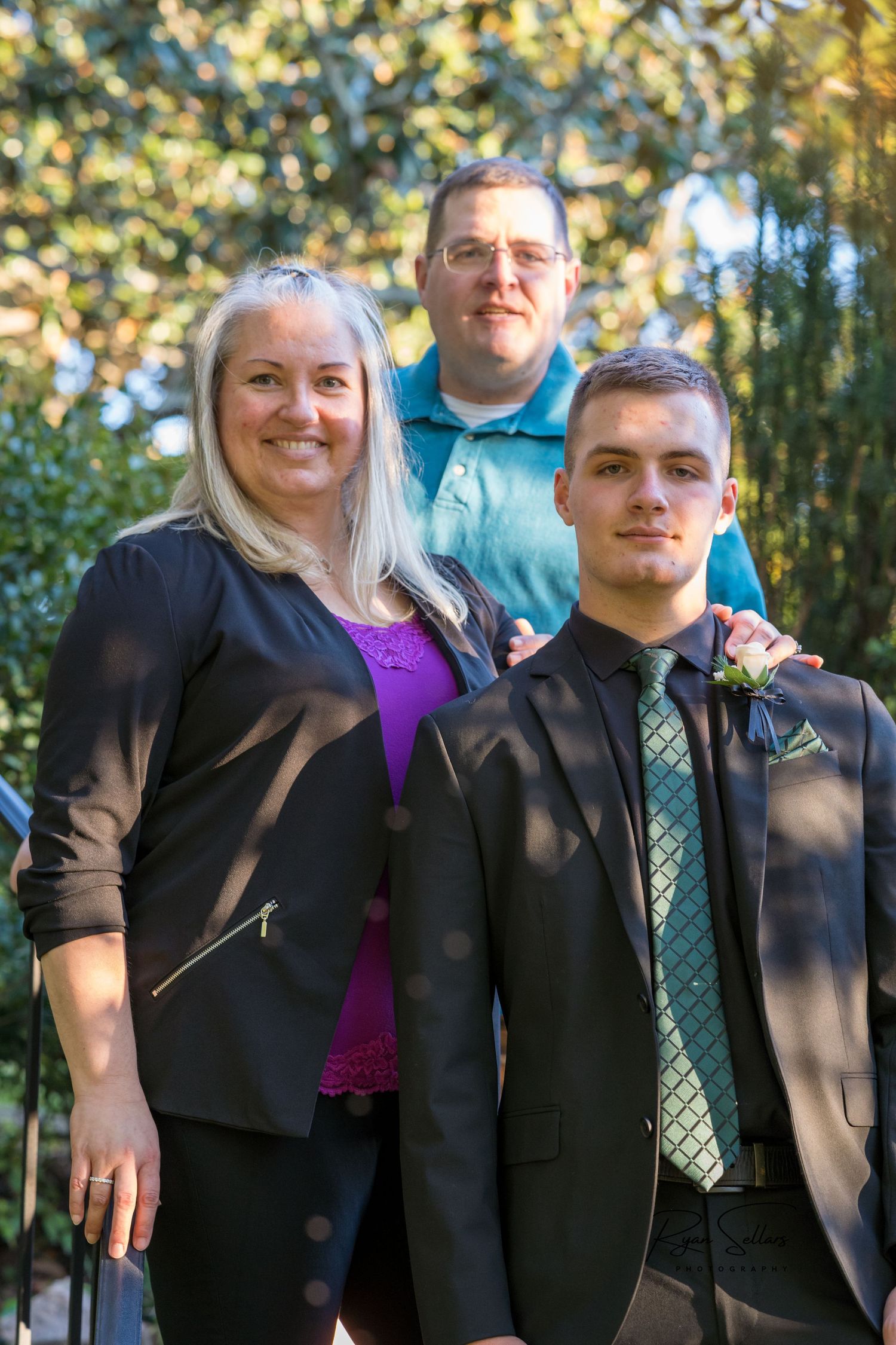 Family poses together outdoors with trees and natural lighting in the background.