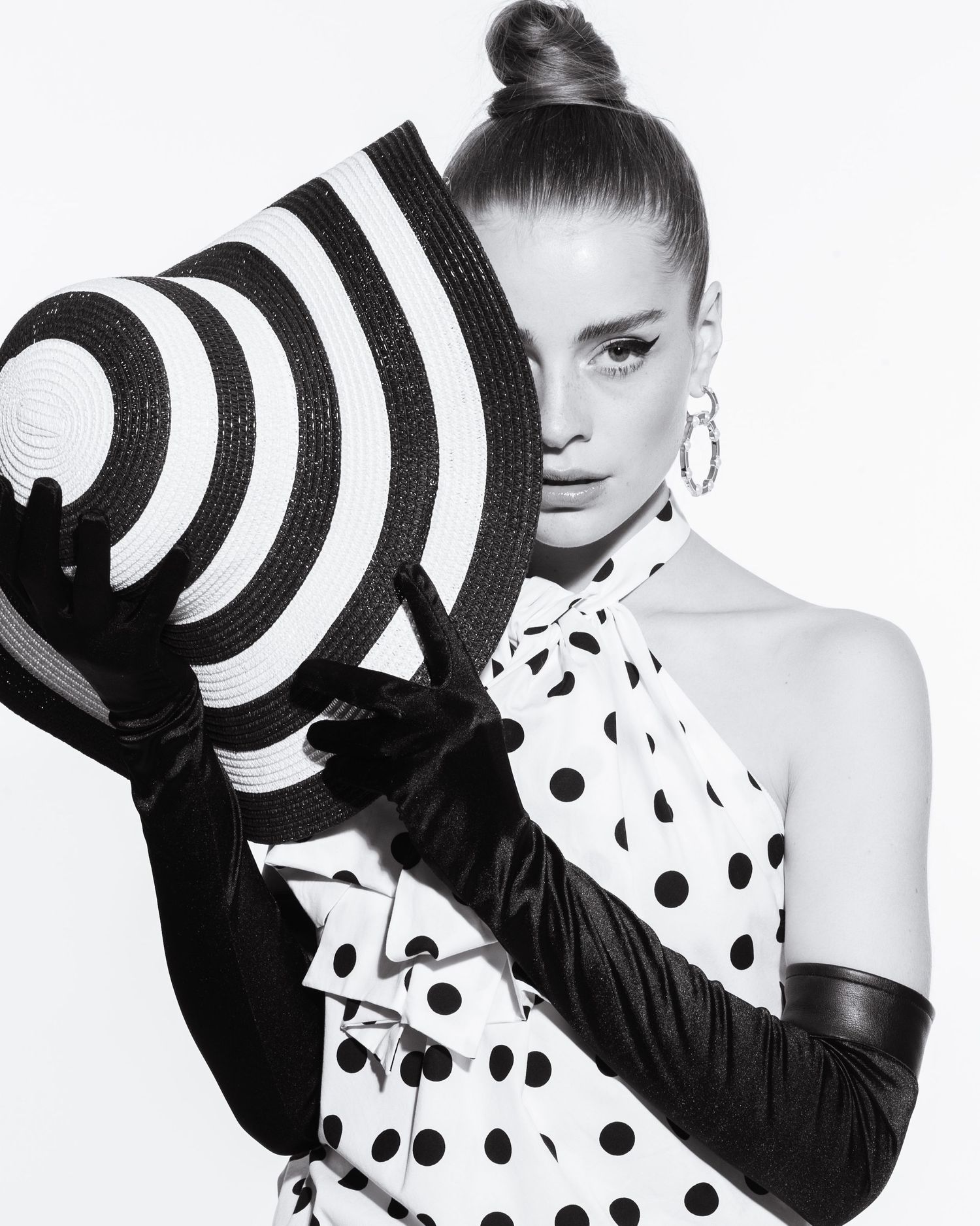 A model in a polka dot dress holds a striped hat with black gloves in a black and white photo.