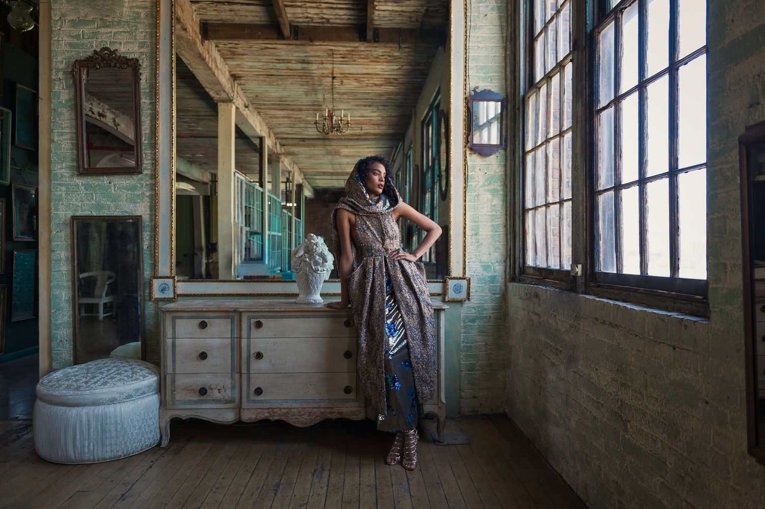 An elegant woman in a patterned dress poses in a rustic vintage hallway with antique furniture.