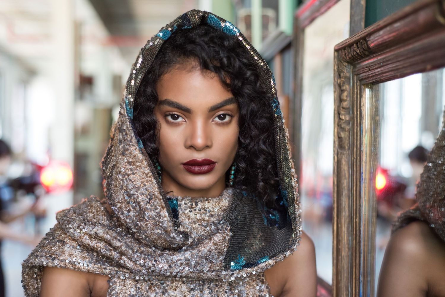 A woman in a glittering hooded wrap poses near a mirror with bold makeup and curly hair.