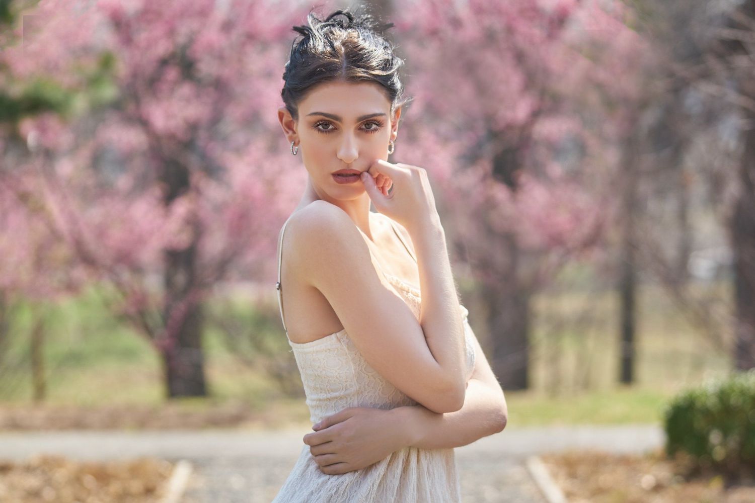 A young woman in a white dress poses gracefully outdoors with blooming pink cherry blossom trees in the background.