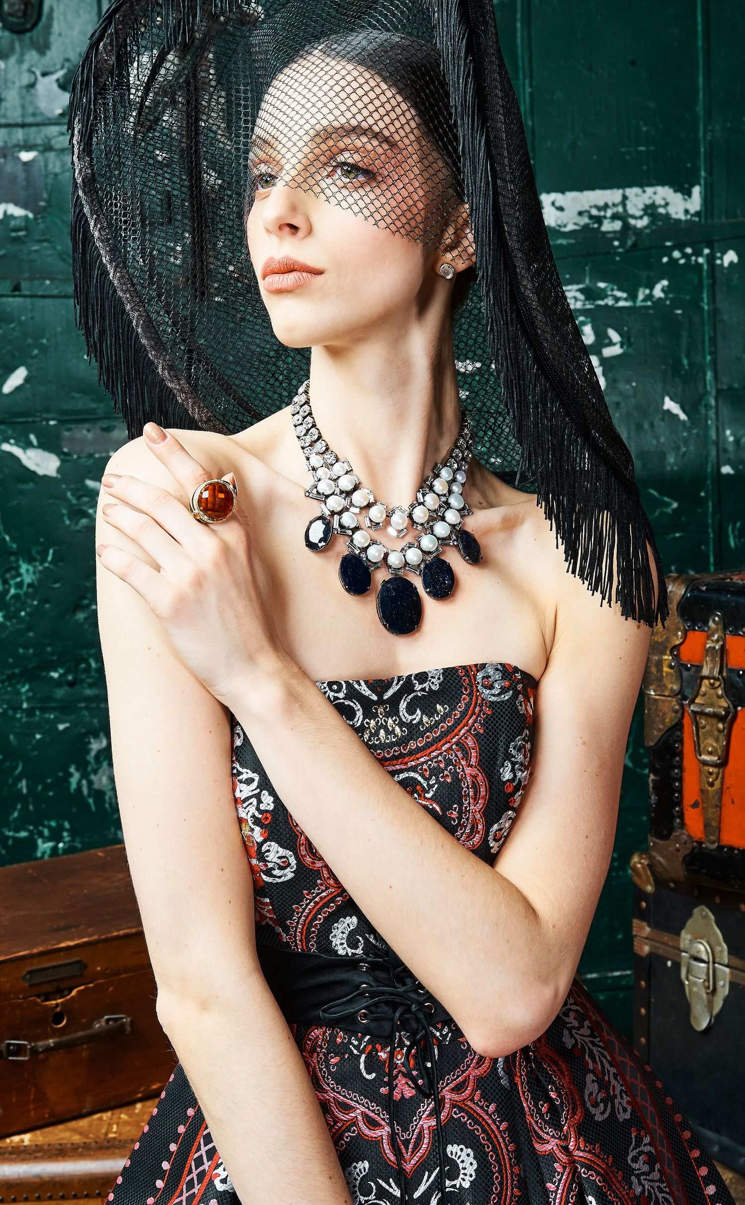 A model in a strapless paisley dress wears a black veil, statement necklace, and amber ring in a vintage setting.