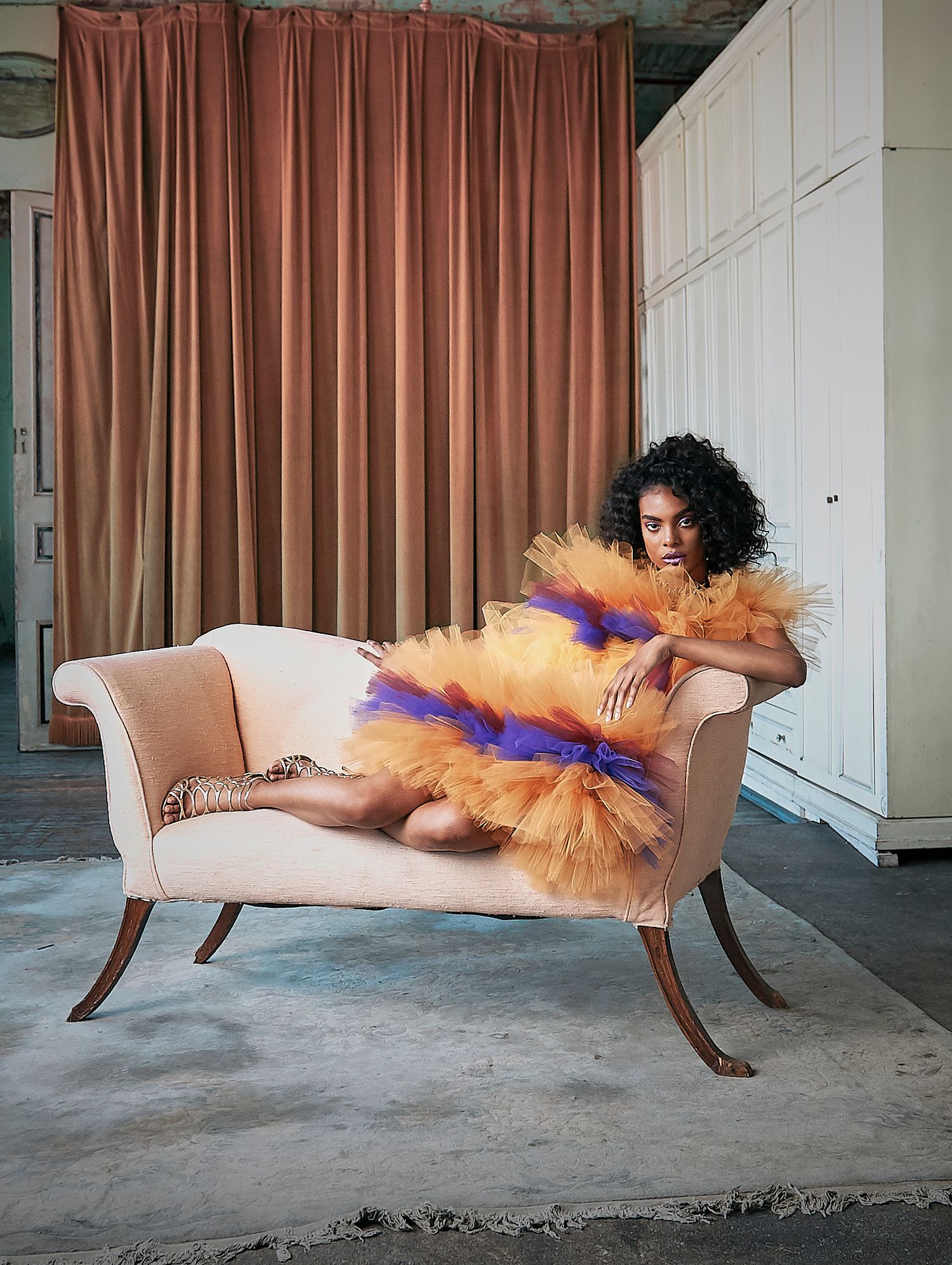 A model in an orange and purple tulle dress reclines on a chaise lounge in an airy room.