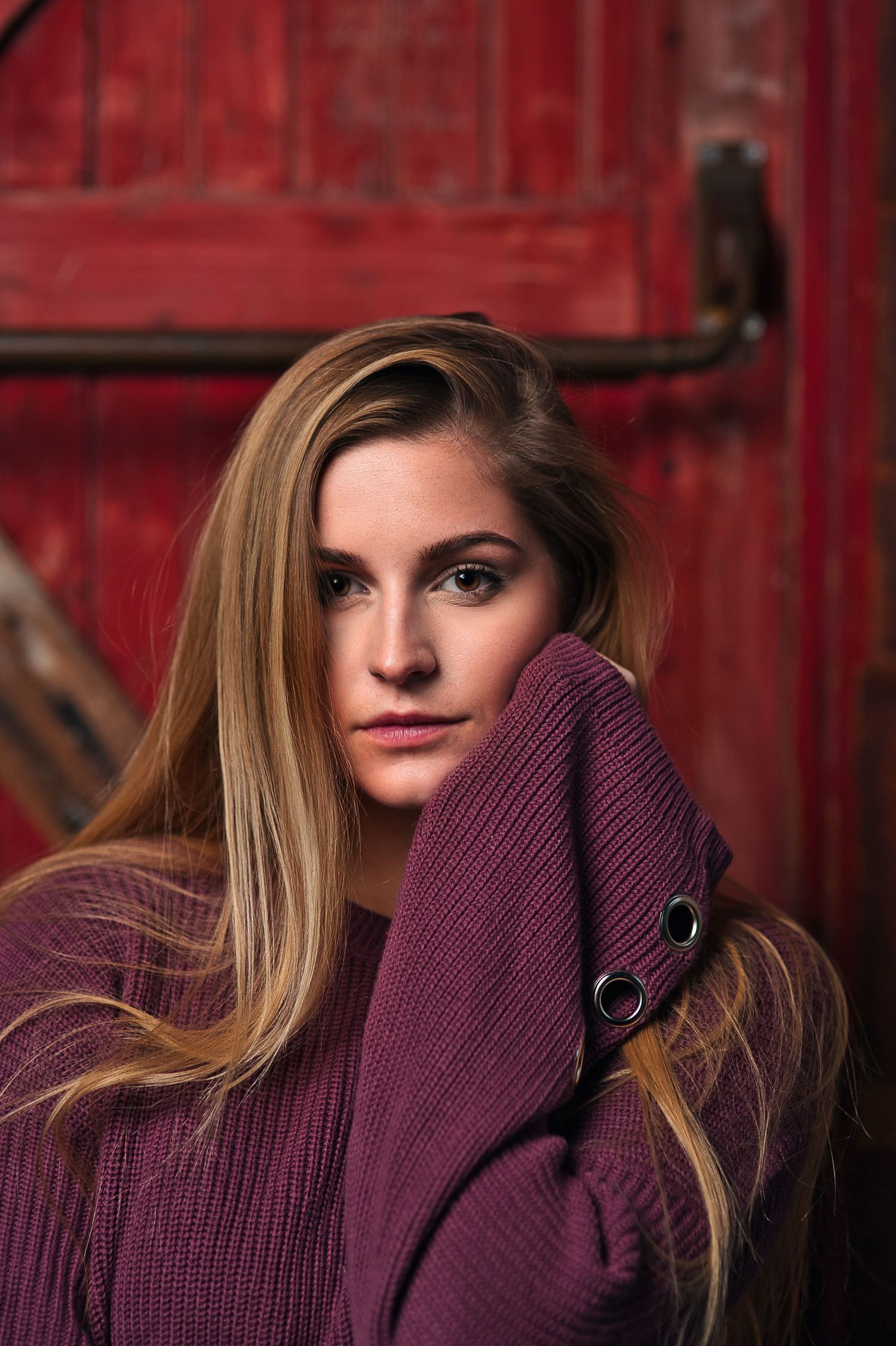 A young woman with long blonde hair wraps herself in a purple knit sweater near a red door.