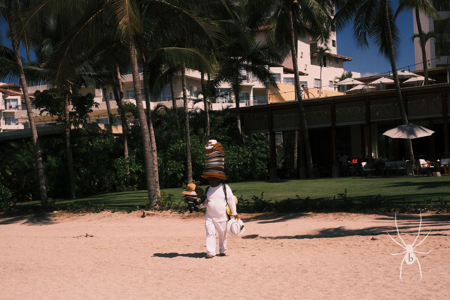 Person in white clothing walking on a beach with palm trees and buildings in background.