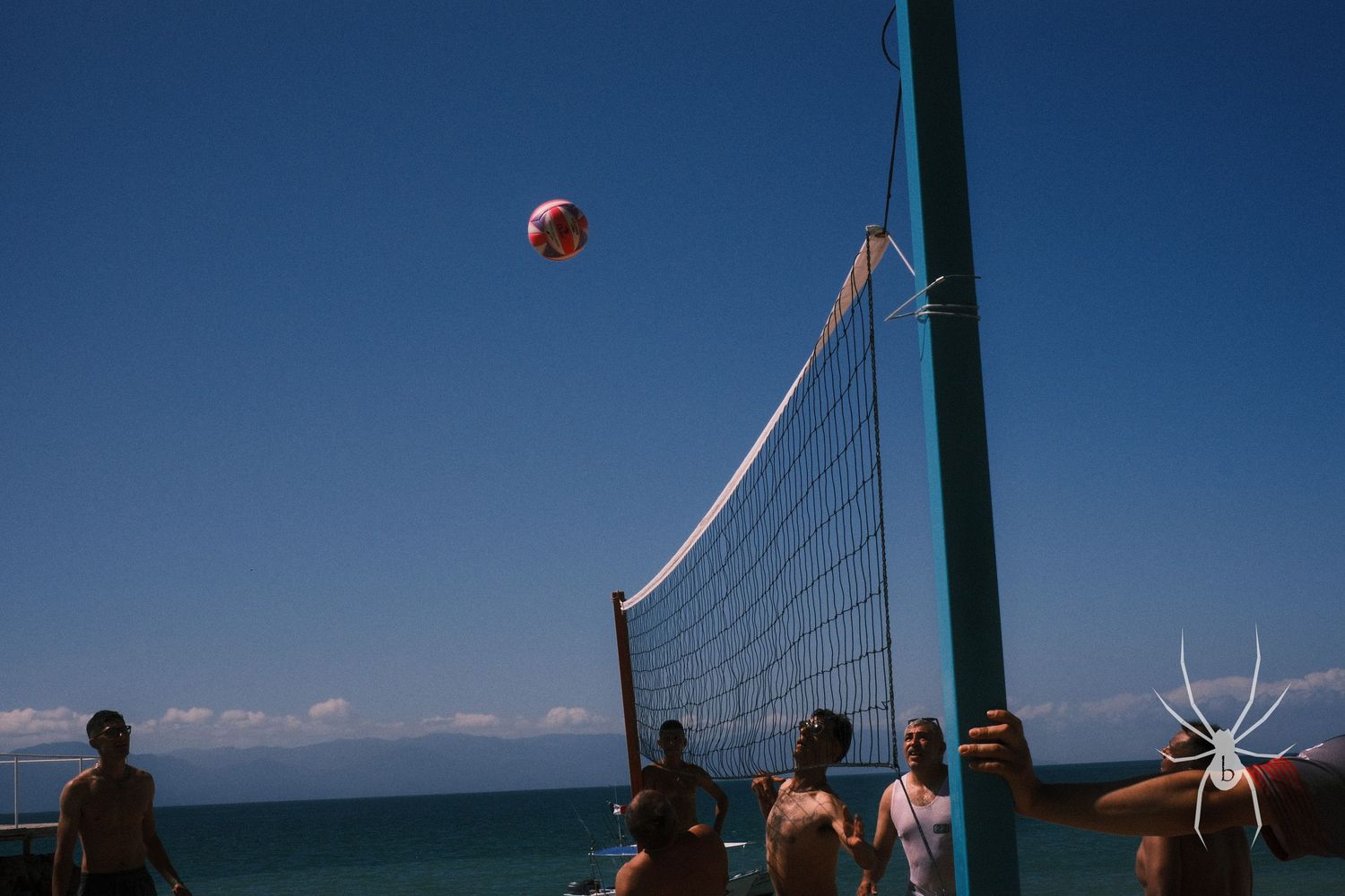 Beach volleyball game in progress against blue sky and ocean backdrop.