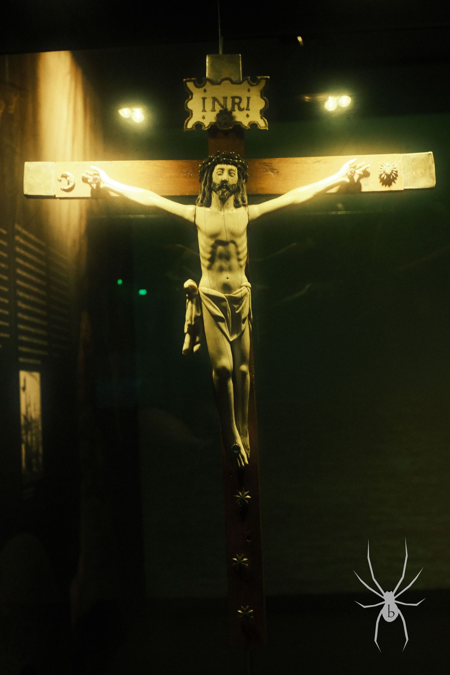 A dark and moody photograph of a crucifix lit from above with INRI inscription and spider motif.