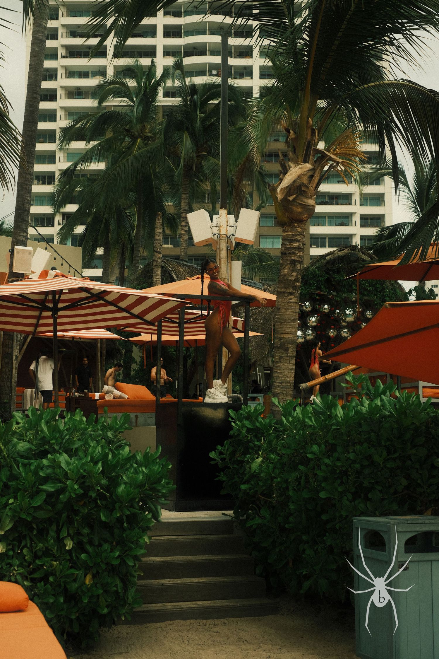 Tropical outdoor dining area with palm trees and orange umbrellas near a high-rise building.