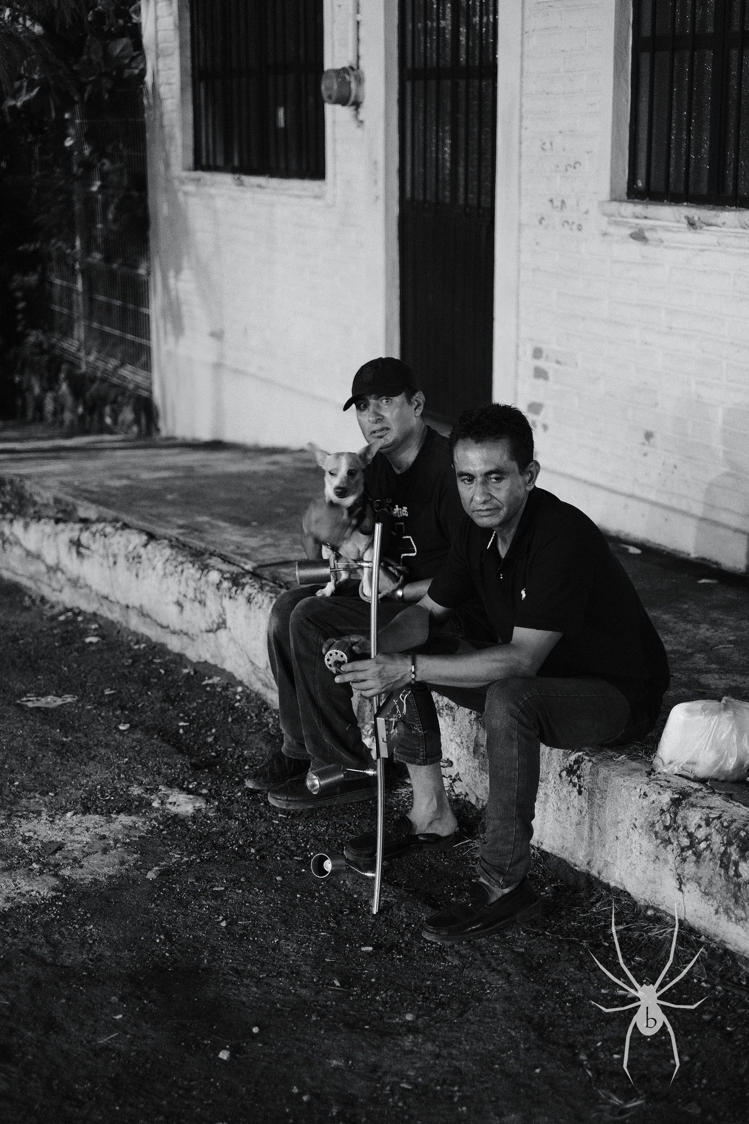 Two figures sit on a concrete ledge at night with a decorative spider element in the corner of the black and white photo.
