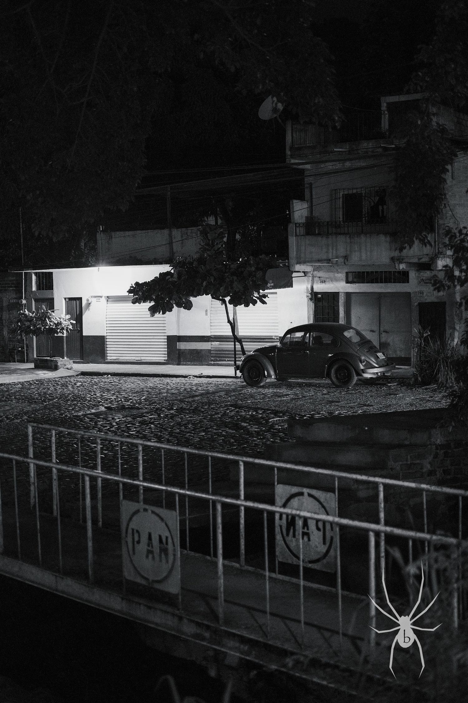 Vintage car parked on dark street with illuminated storefront in black and white.