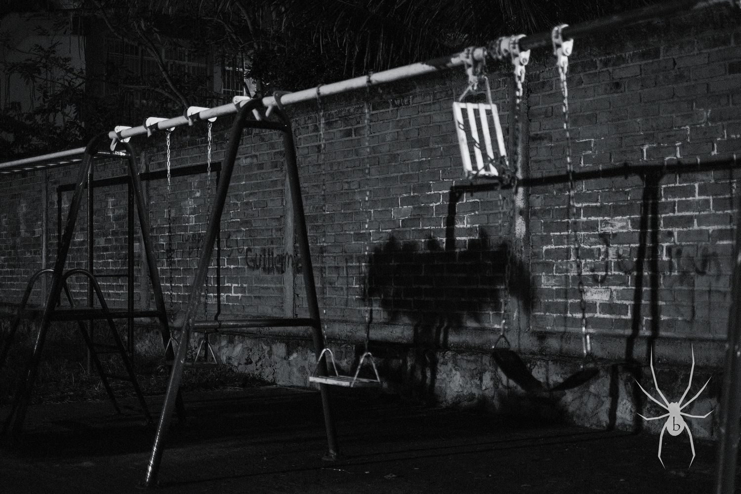 Empty playground swings in black and white with graffiti on brick wall behind.