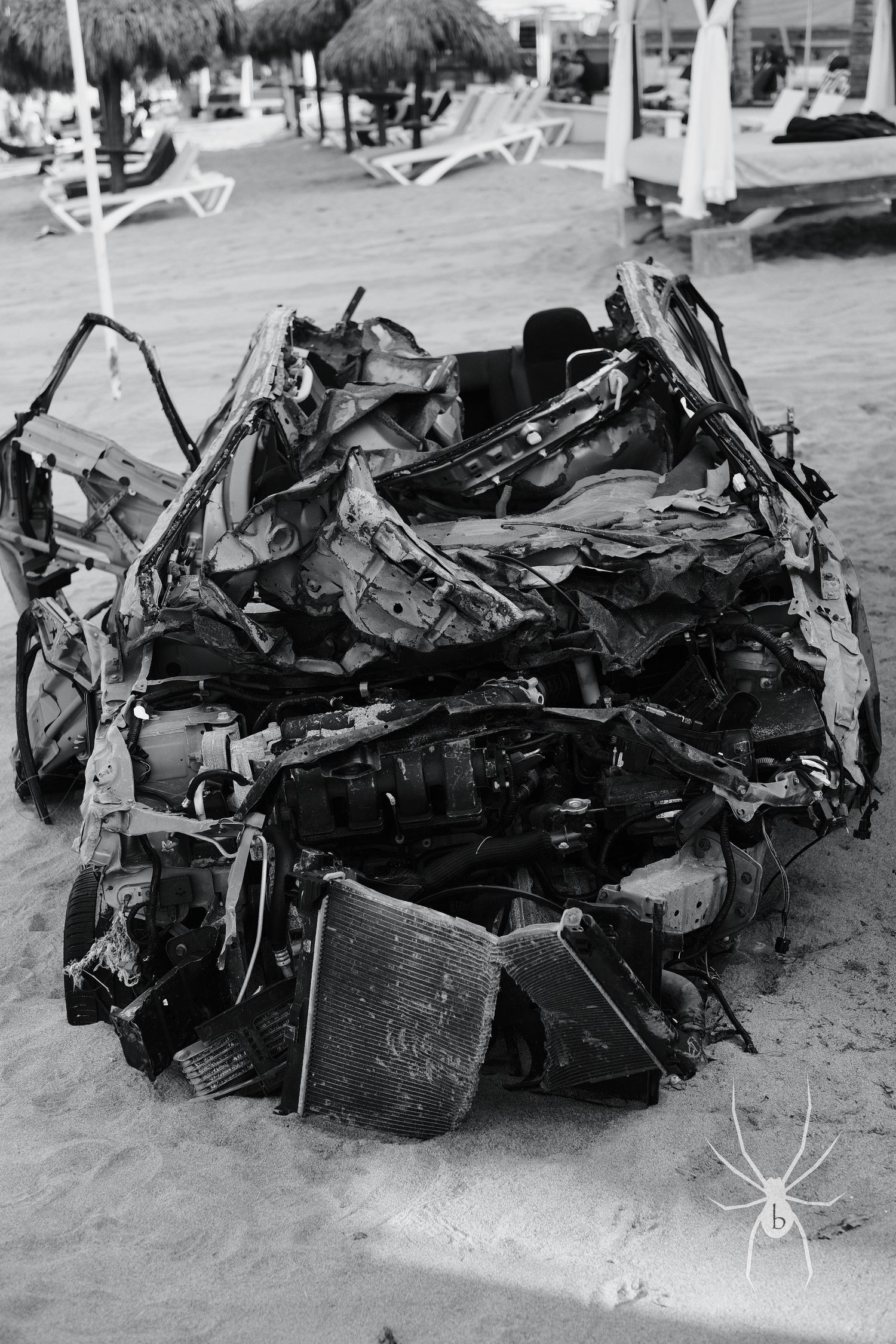 Crushed and mangled car wreckage on sandy beach with lounge chairs in background.